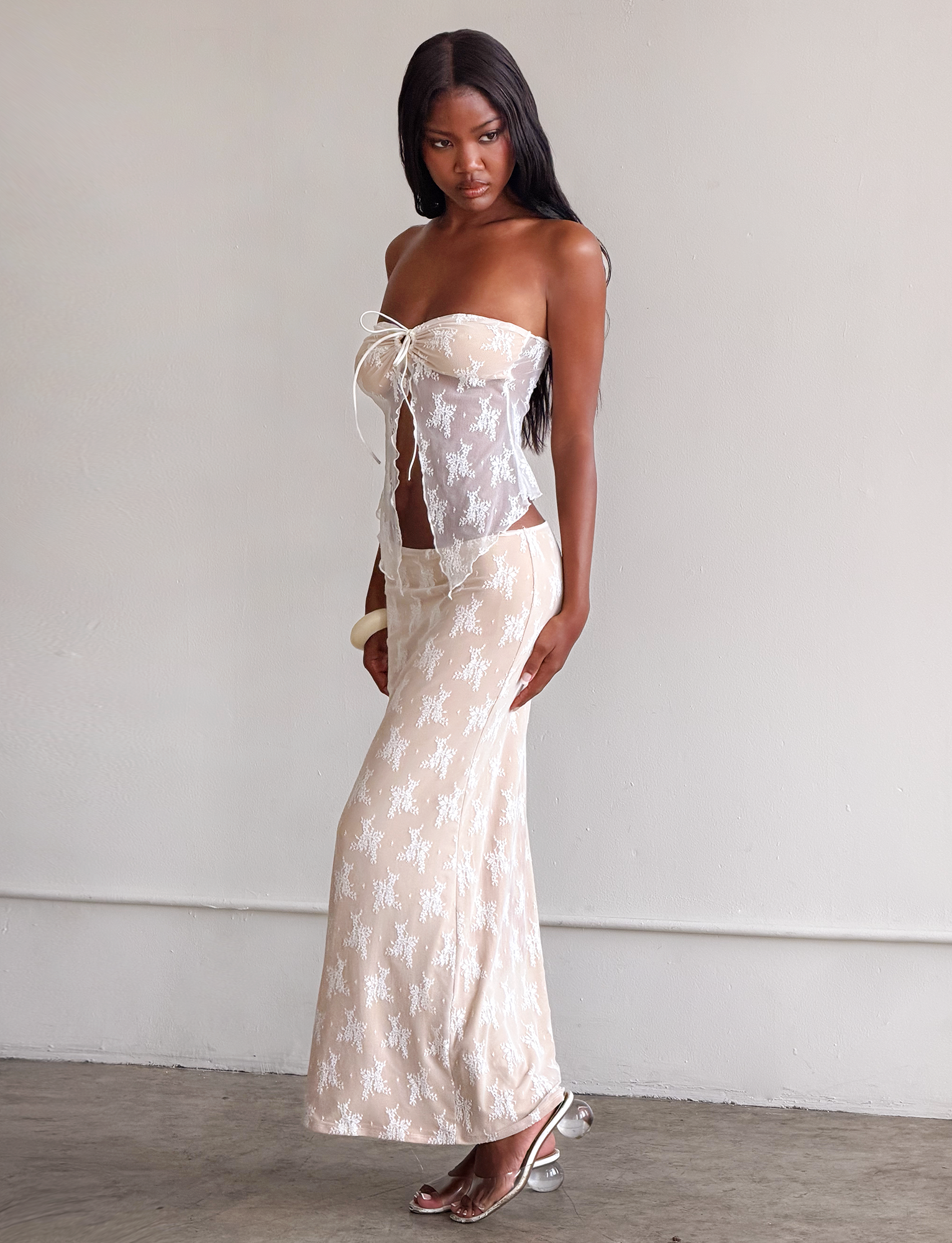A woman stands indoors against a plain light wall, wearing the TIGERMIST KAROLINA TOP - WHITE: a strapless white lace top with front tie and subtle bust cutout, paired with a matching long skirt. She has long hair and open-toe heels.