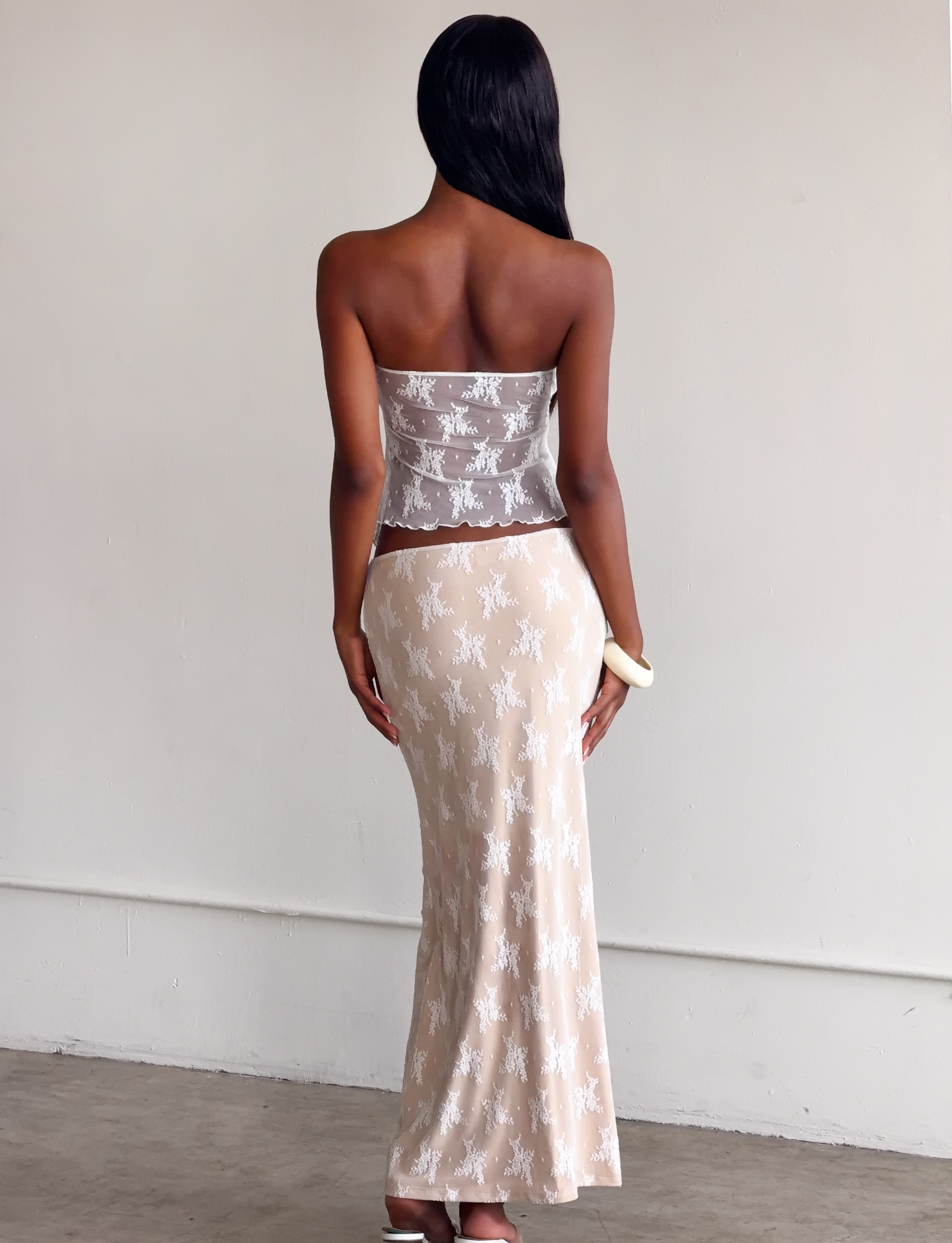 A woman with long dark hair, seen from behind, wears a strapless sheer top and the TIGERMIST KAROLINA SKIRT - WHITE LACE with white floral patterns, holding a small white clutch in her right hand.
