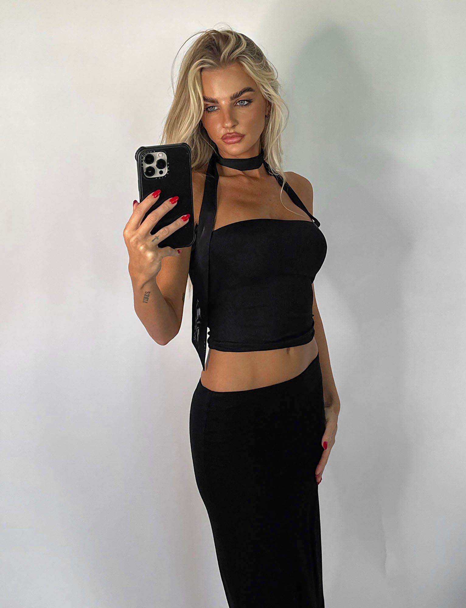 A person with long blonde hair is taking a mirror selfie with a smartphone. They are wearing the CHRYSLER TOP - BLACK and a black skirt from TIGERMIST. The background is plain white.