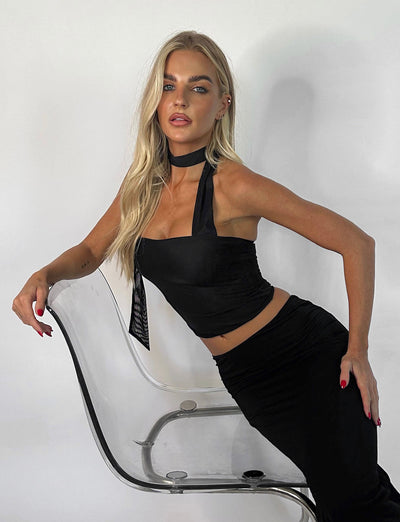 A woman with long blonde hair leans against a clear, modern chair, showcasing her sleek TIGERMIST CHRYSLER TOP - BLACK and matching skirt. She has a black choker around her neck, and her nails are painted red. She gazes directly at the camera against a plain white background.