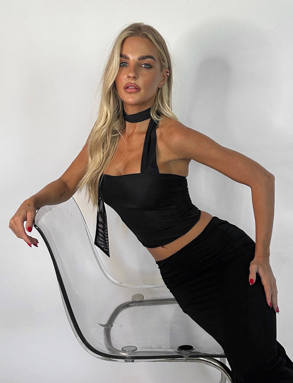 A woman with long blonde hair leans against a clear, modern chair, showcasing her sleek TIGERMIST CHRYSLER TOP - BLACK and matching skirt. She has a black choker around her neck, and her nails are painted red. She gazes directly at the camera against a plain white background.