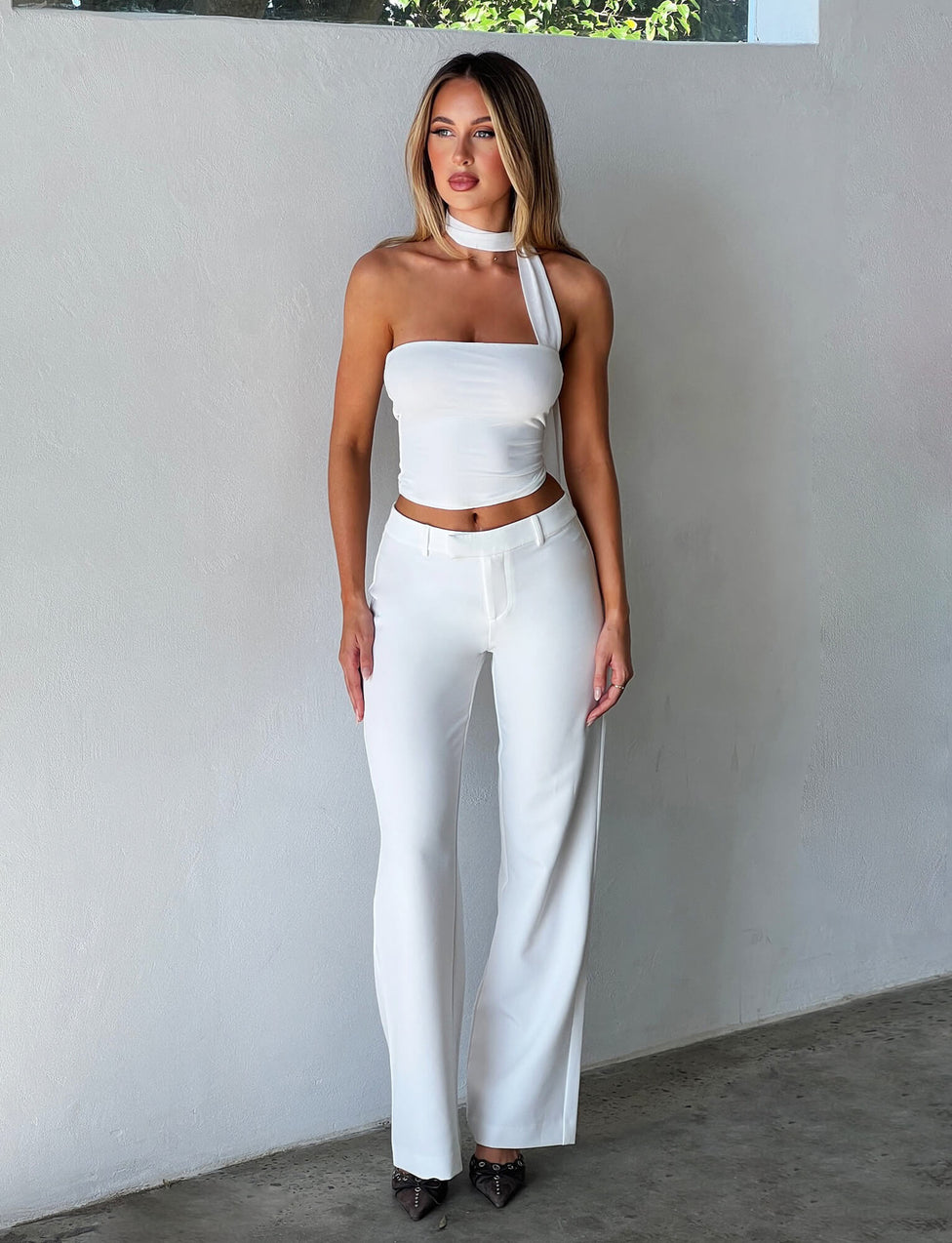 A woman stands against a plain white background, wearing an all-white TIGERMIST ensemble featuring the CHRYSLER TOP - WHITE, which has a unique neck strap. She pairs the sleeveless top with high-waisted, wide-leg trousers. Her long, straight hair complements her pointed black shoes.