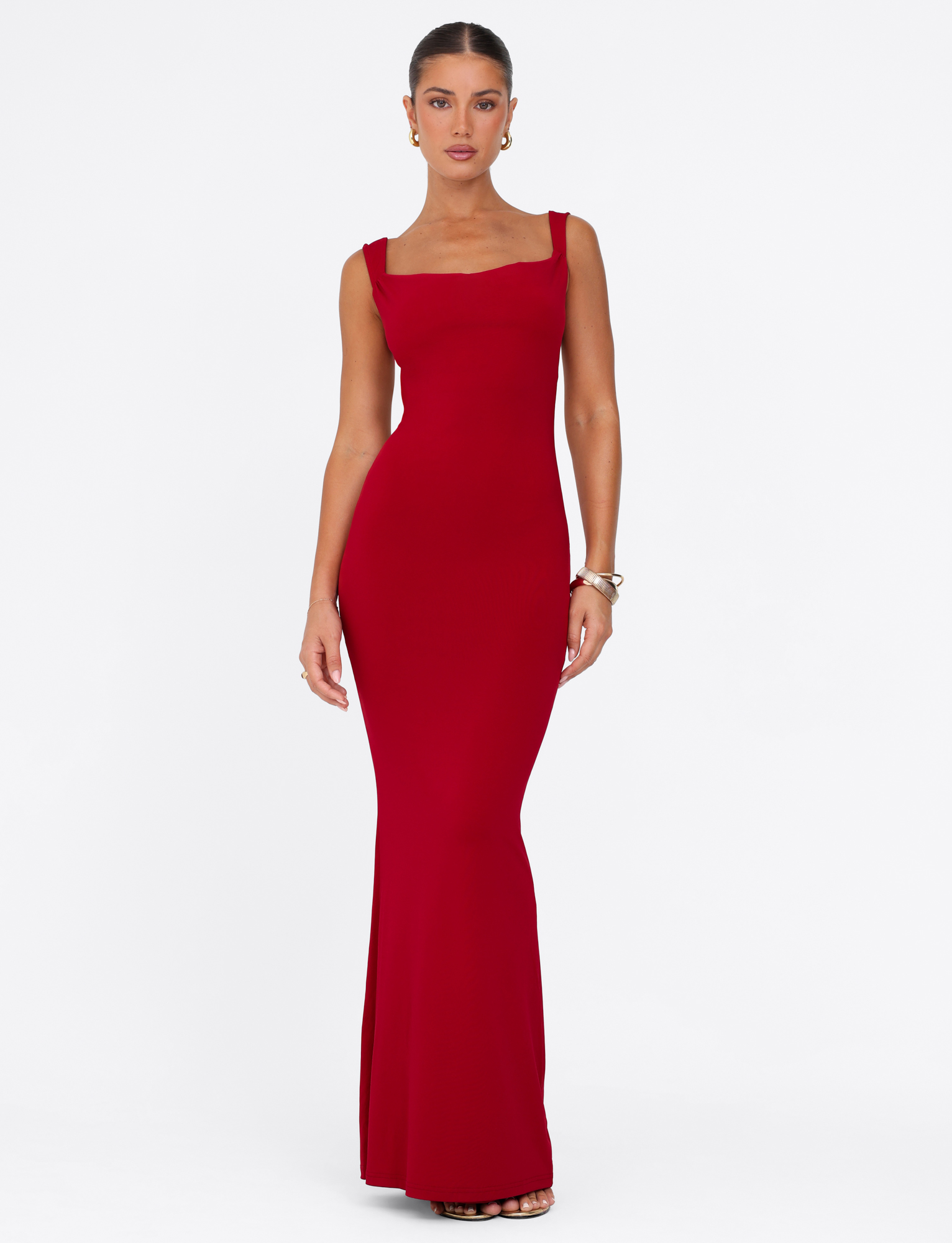 A woman models the TIGERMIST MORANA MAXI DRESS - RED, a fitted floor-length dress with wide straps, against a white background. Her hair is pulled back and she wears earrings, bracelets, and open-toe heels.
