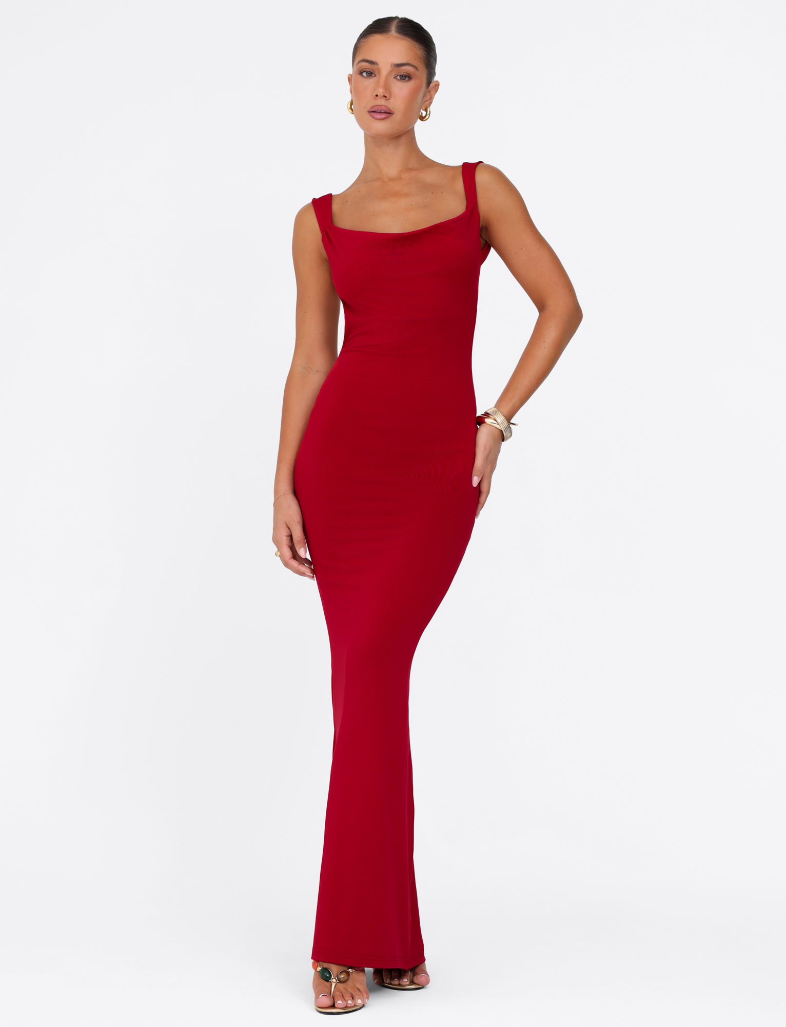 A woman poses against a white background in the TIGERMIST MORANA MAXI DRESS - RED, a sleeveless, floor-length bodycon dress with a square neckline. She styles it with pulled-back hair, earrings, bracelets, and open-toe sandals.