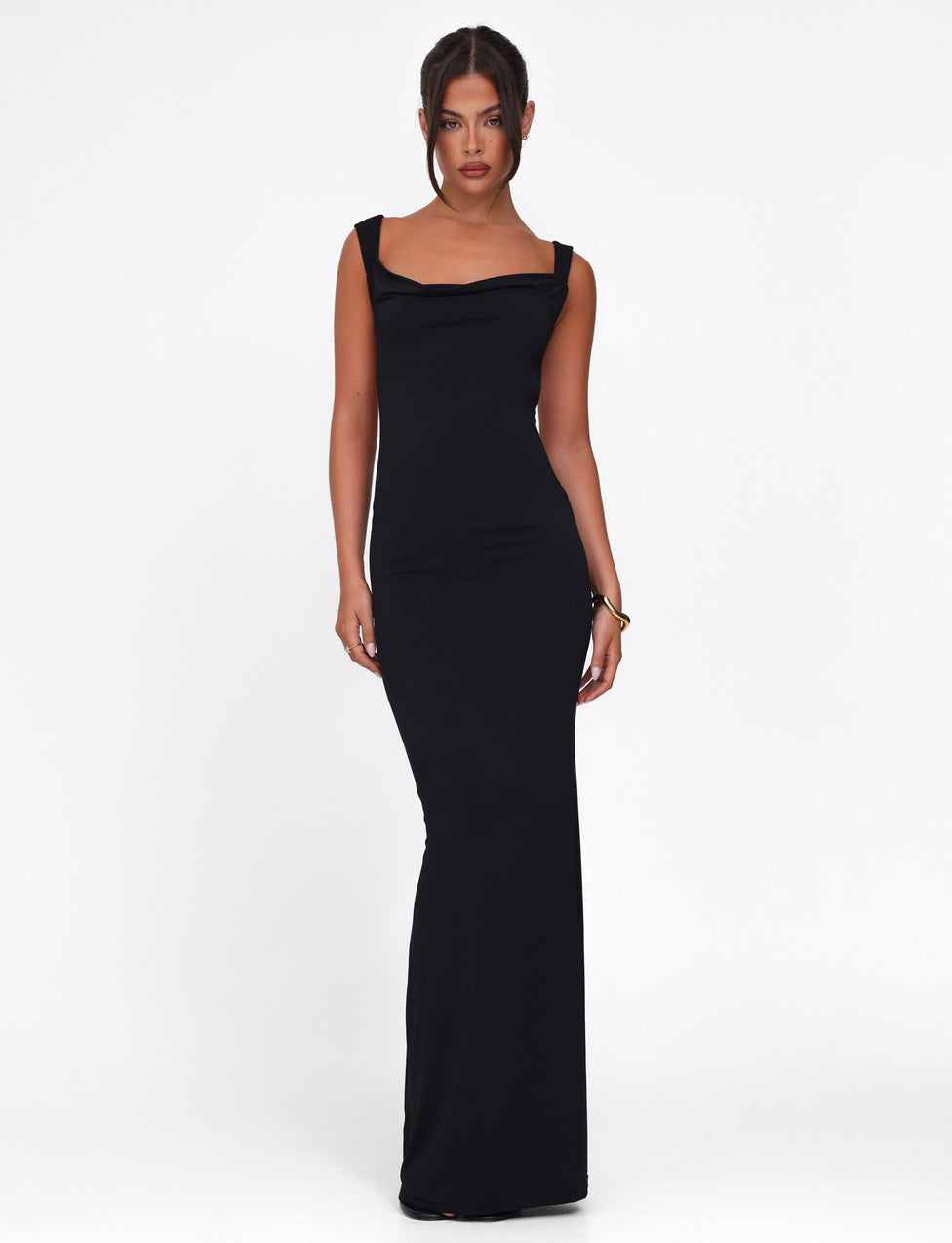 In a sleek Morana Dress by Tigermist, they pose confidently. The black dress features a long, elegant bodycon fit with thick shoulder straps. Their dark hair is neatly styled, and a bracelet adorns their right wrist, set against a plain white background.
