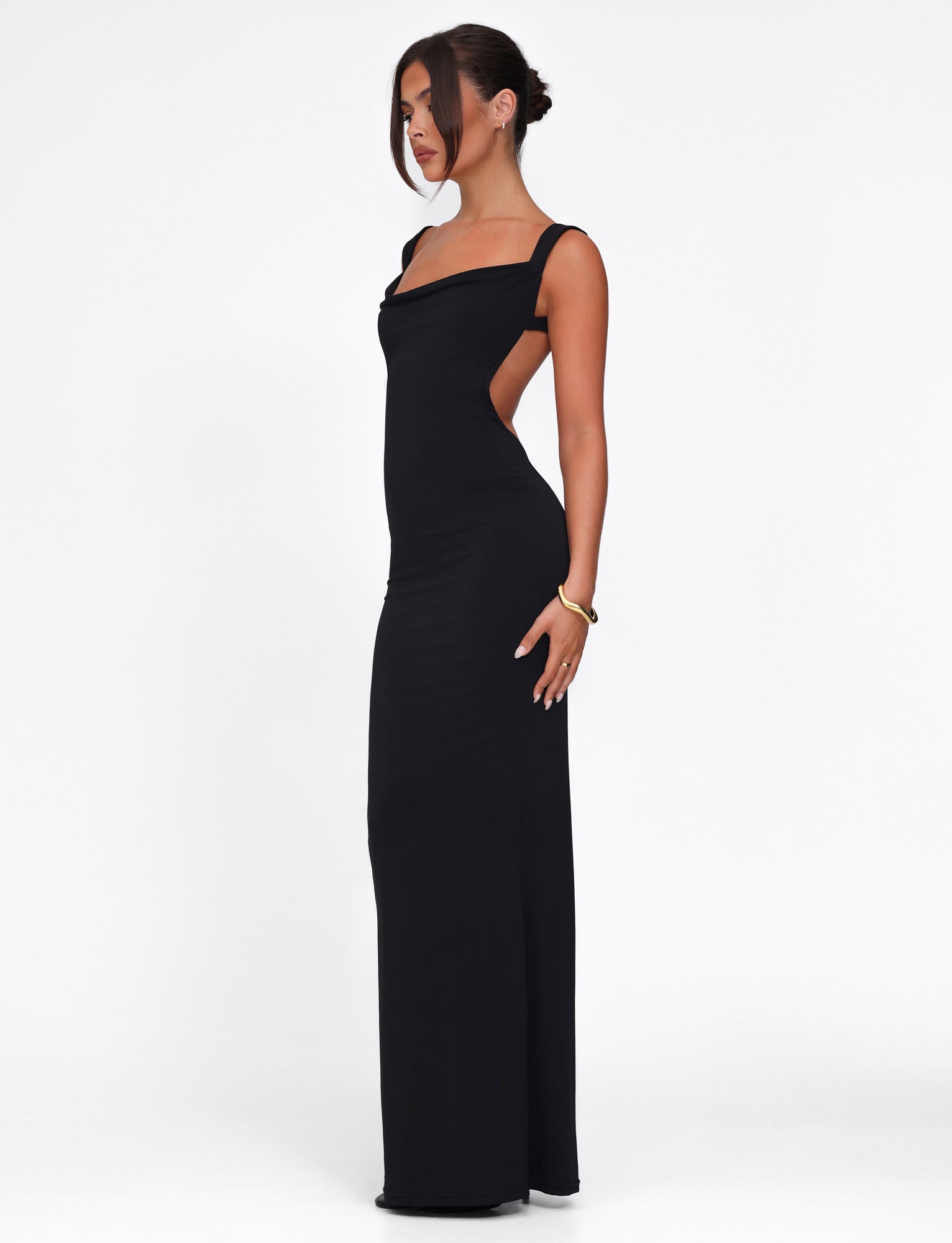 A woman models the form-fitting MORANA DRESS - BLACK by TIGERMIST against a white background, highlighting its open back and square neckline. Her updo hairstyle complements the elegant bracelet on her left wrist.