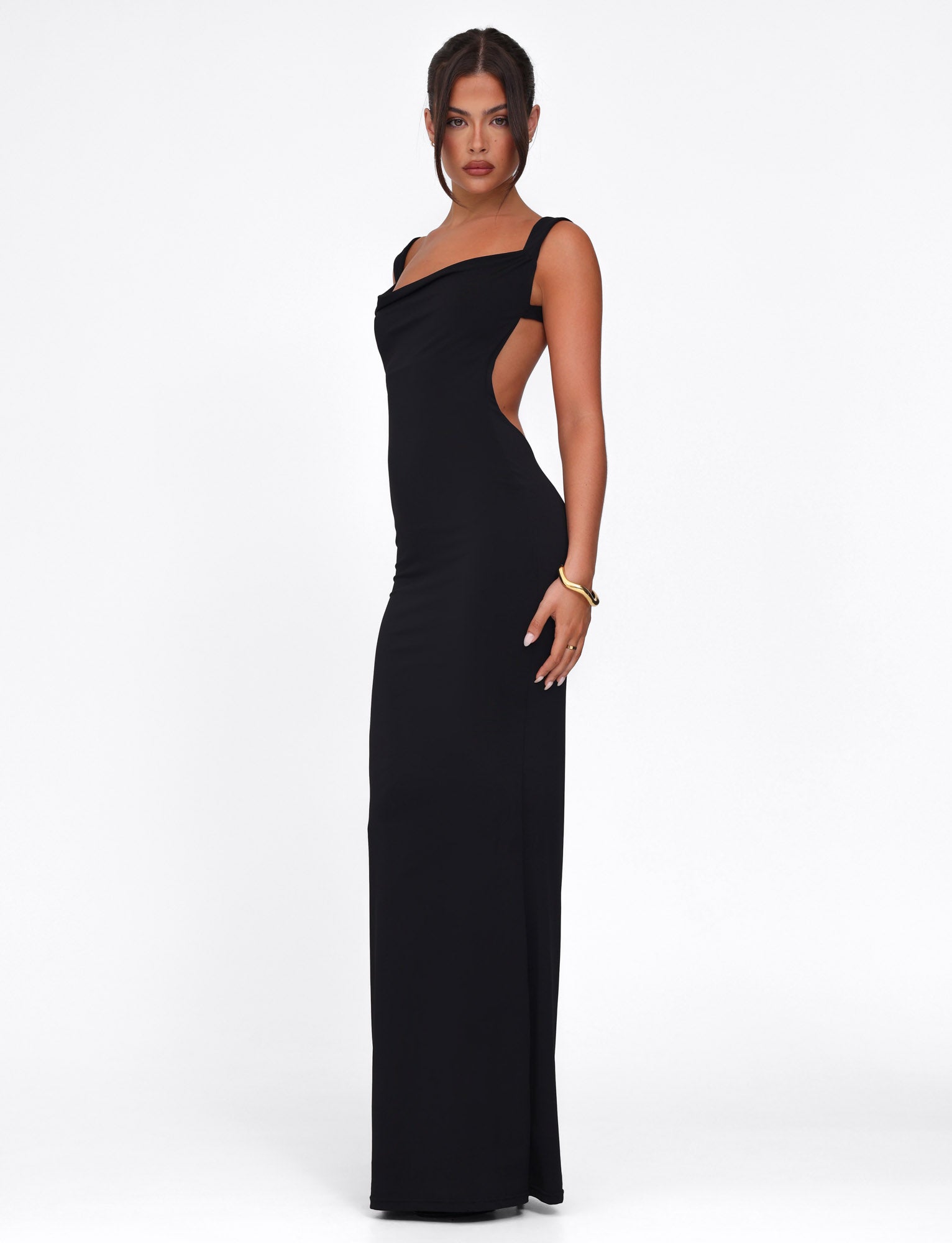 A woman poses against a white background in the sleek TIGERMIST MORANA DRESS - BLACK. The asymmetrical neckline and open sides of this bodycon design enhance her elegance. She wears a single gold bracelet with her hair in an updo, complementing the minimalist allure.