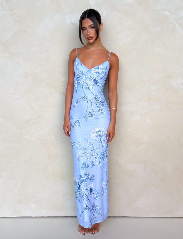 A woman poses against a textured beige wall in the TIGERMIST JULIANNA DRESS – BLUE: CERAMIC FLORAL, featuring thin straps and a V-neckline. Her hair is styled up, and she pairs the look with clear-heeled shoes.