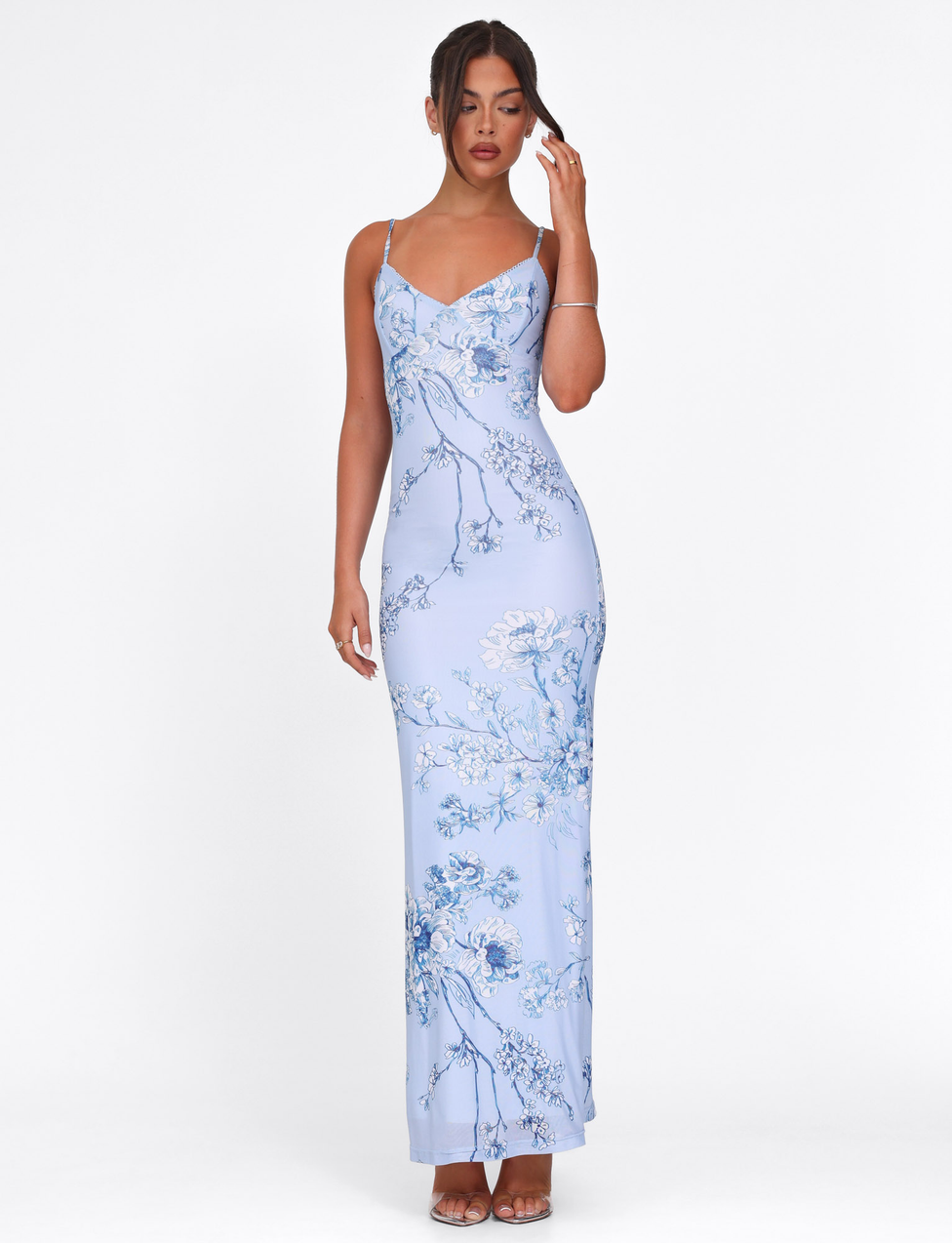 A woman poses against a white background wearing the TIGERMIST JULIANNA DRESS in Blue Ceramic Floral, a floor-length maxi with thin straps and a V-neckline. Her hair is up; one hand is near her face and the other rests by her side.