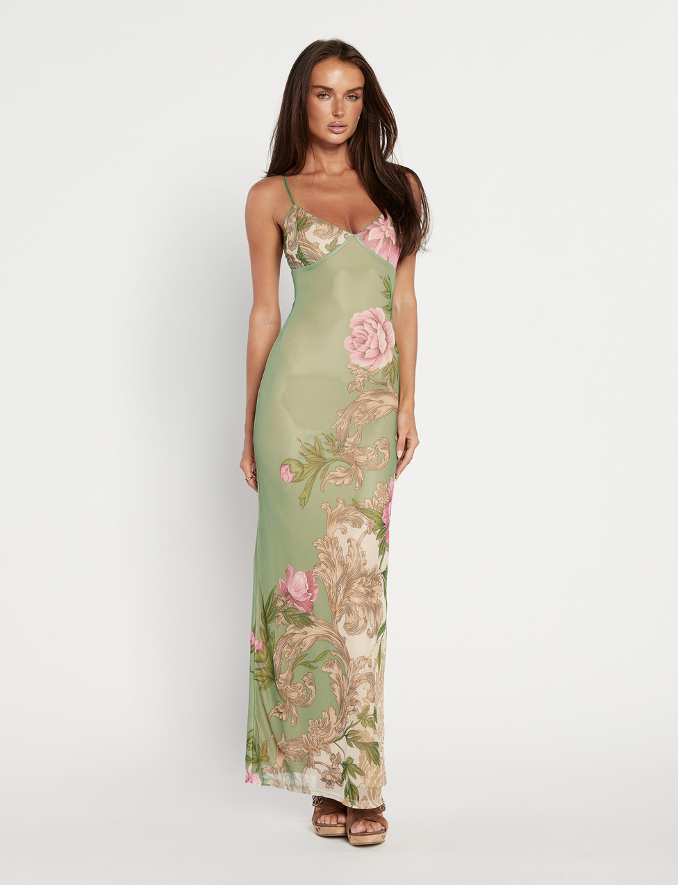 A woman with long brown hair and a neutral expression wears the TIGERMIST JULIANNA DRESS in Green Roccoco Floral, featuring adjustable spaghetti straps, pink floral and ornate beige patterns, paired with brown platform sandals against a white background.