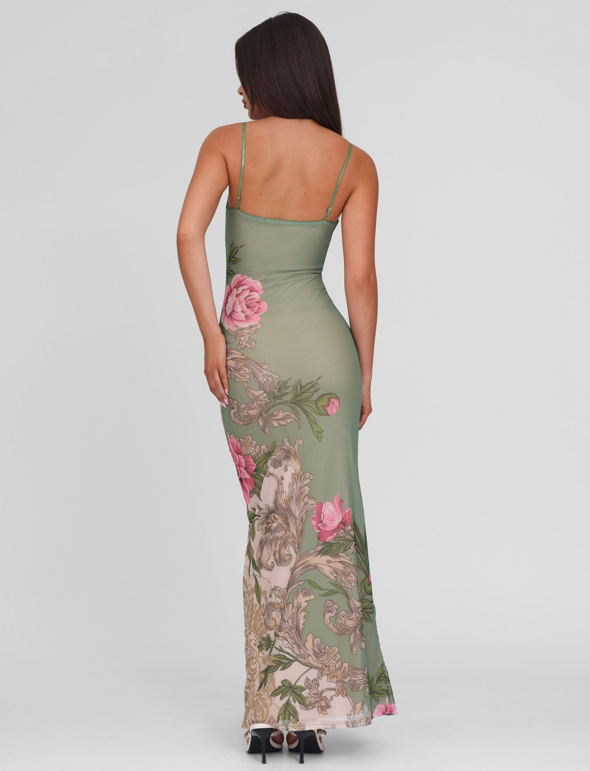 A woman with long dark hair faces away, wearing the TIGERMIST JULIANNA DRESS in green Roccoco floral—a sleeveless maxi with pink flowers, ornate hem, and thin straps—paired stylishly with high heels.