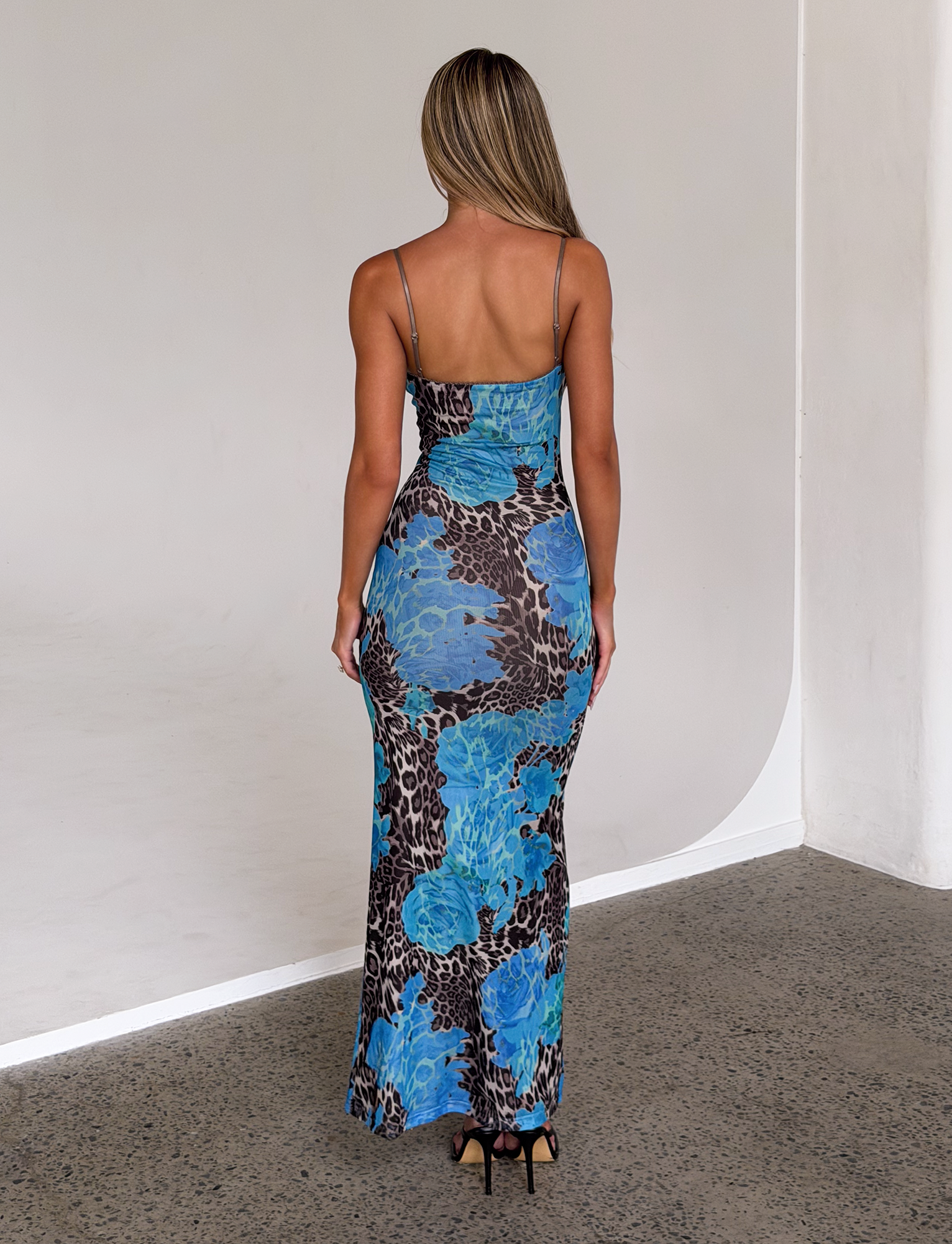 A woman with long, light brown hair stands indoors on a polished concrete floor, wearing the TIGERMIST JULIANNA DRESS - BLUE : LEOPARD and black high heels.