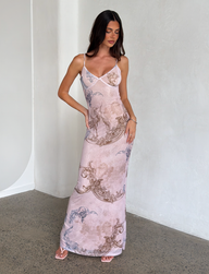 A woman stands indoors against a light wall, wearing the TIGERMIST JULIANNA DRESS - MULTI: COLLAGE: CRANES, a long maxi dress with intricate crane and collage prints. She has long dark hair, strappy heels, and poses elegantly with one hand on her hip.