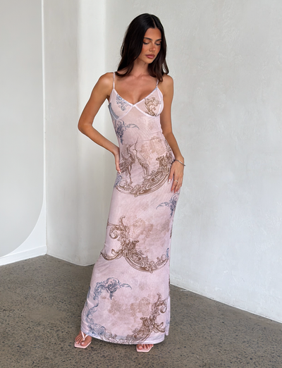 A woman stands indoors against a light wall, wearing the TIGERMIST JULIANNA DRESS - MULTI: COLLAGE: CRANES, a long maxi dress with intricate crane and collage prints. She has long dark hair, strappy heels, and poses elegantly with one hand on her hip.