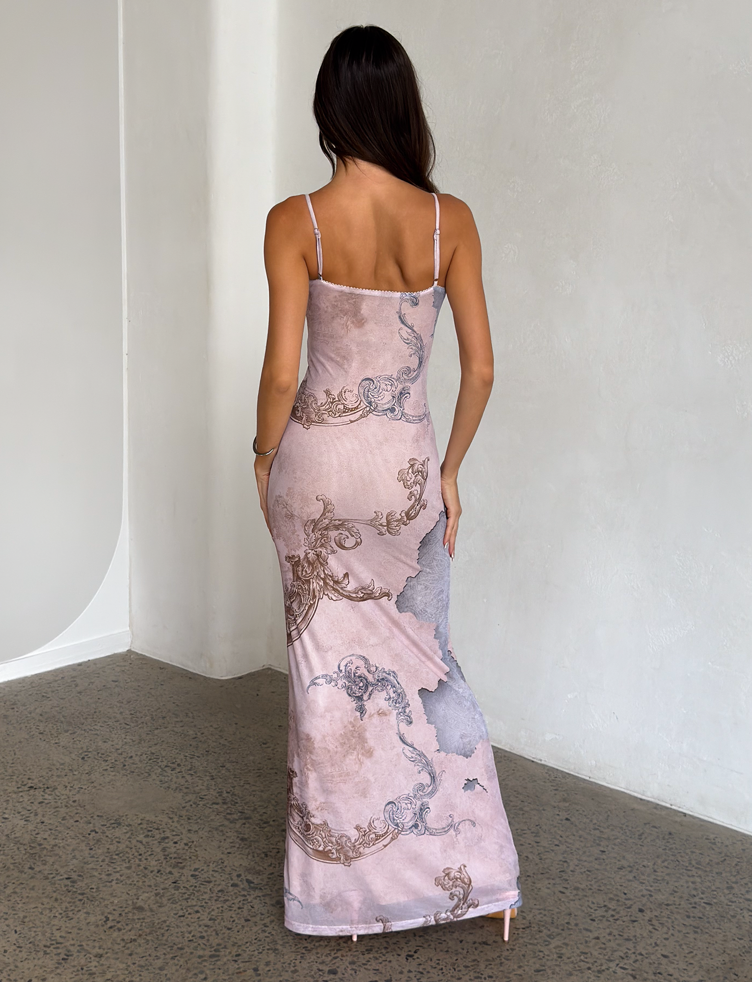 A woman with long dark hair stands indoors facing a white textured wall, wearing the sleeveless, floor-length JULIANNA DRESS - MULTI : COLLAGE : CRANES by TIGERMIST, which features an intricate swirling pattern.