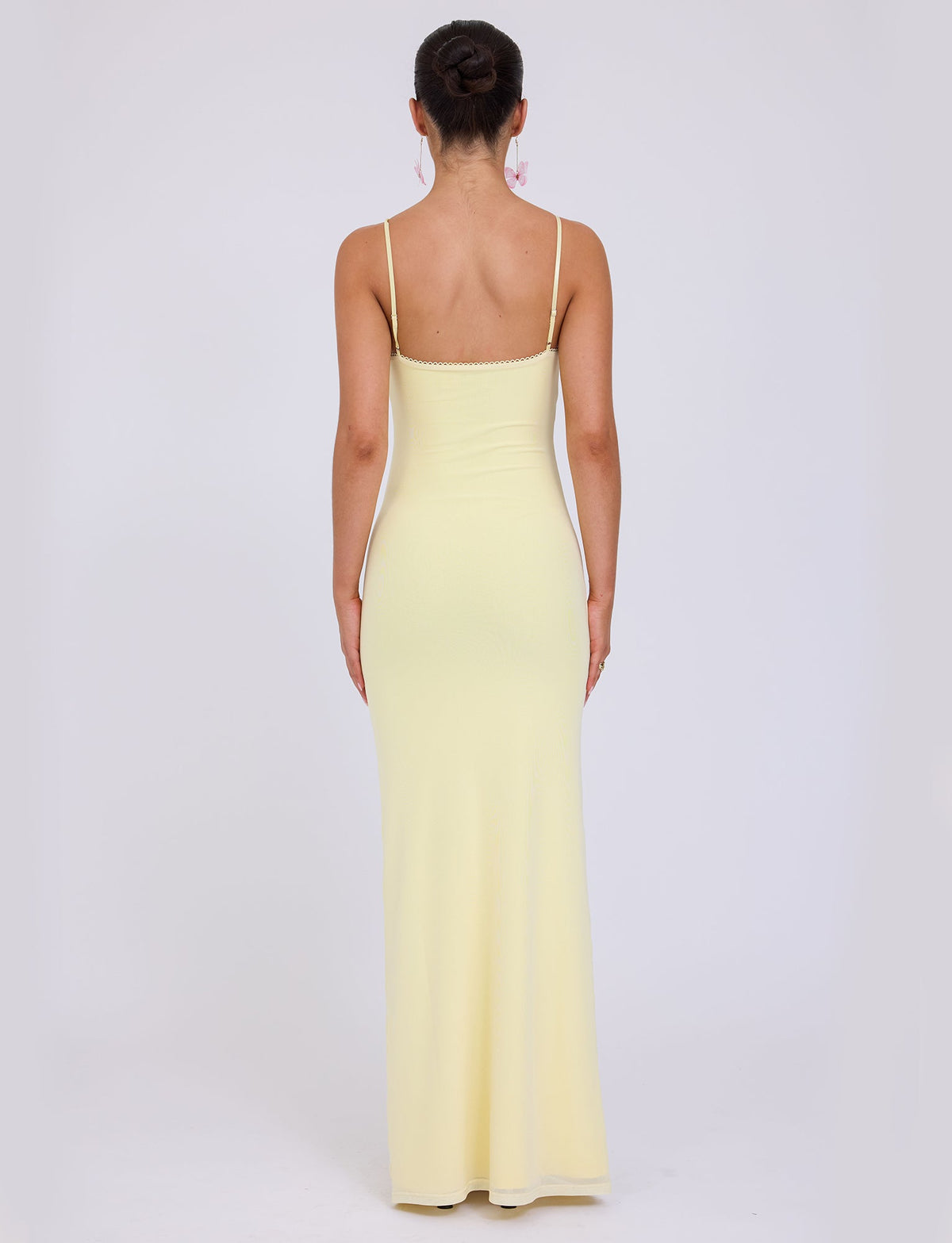 A woman stands with her back to the camera, wearing the JULIANNA DRESS - YELLOW : LEMON by TIGERMIST, a long, fitted dress with thin shoulder straps. Her dark hair is styled in a high bun, and she accessorizes with dangling earrings. The background is plain white.
