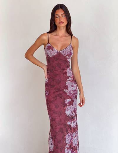 A woman models the TIGERMIST JULIANNA DRESS - MULTI : FLORAL : ROSE STRIPE, a fitted, sleeveless dress with pink floral stripe print, standing by a light textured wall with one hand on her hip and looking slightly to the left.