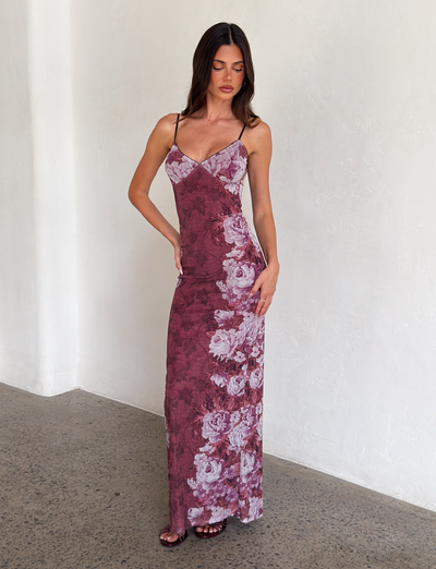 A woman with long brown hair stands indoors by a white textured wall, wearing the TIGERMIST JULIANNA DRESS - MULTI: a sleeveless, floor-length maxi with thin straps and a rose stripe floral print. She looks down.
