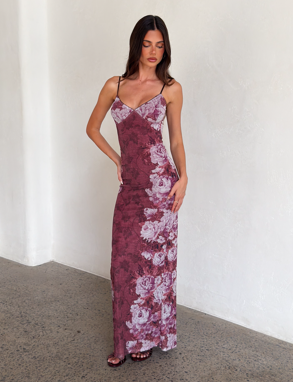 A woman with long brown hair stands indoors by a white textured wall, wearing the TIGERMIST JULIANNA DRESS - MULTI: a sleeveless, floor-length maxi with thin straps and a rose stripe floral print. She looks down.