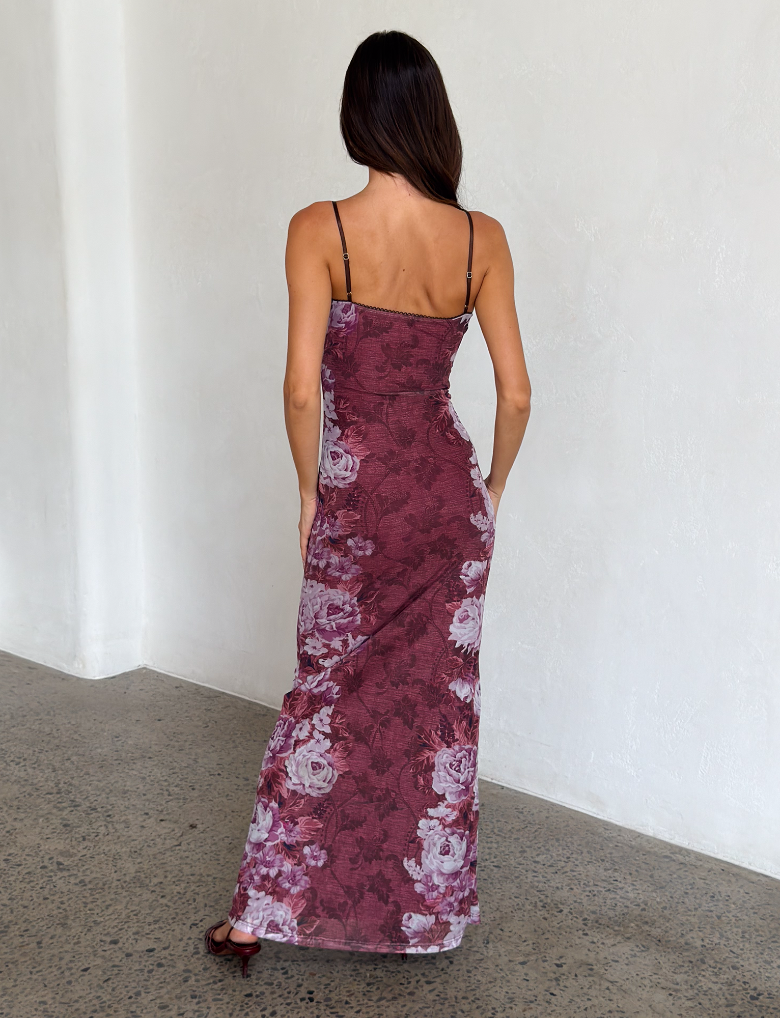 A woman with long brown hair faces away, wearing the TIGERMIST JULIANNA DRESS— a sleeveless, floor-length maxi with thin straps and a multi floral rose stripe print. She stands on a speckled floor against a plain white wall.