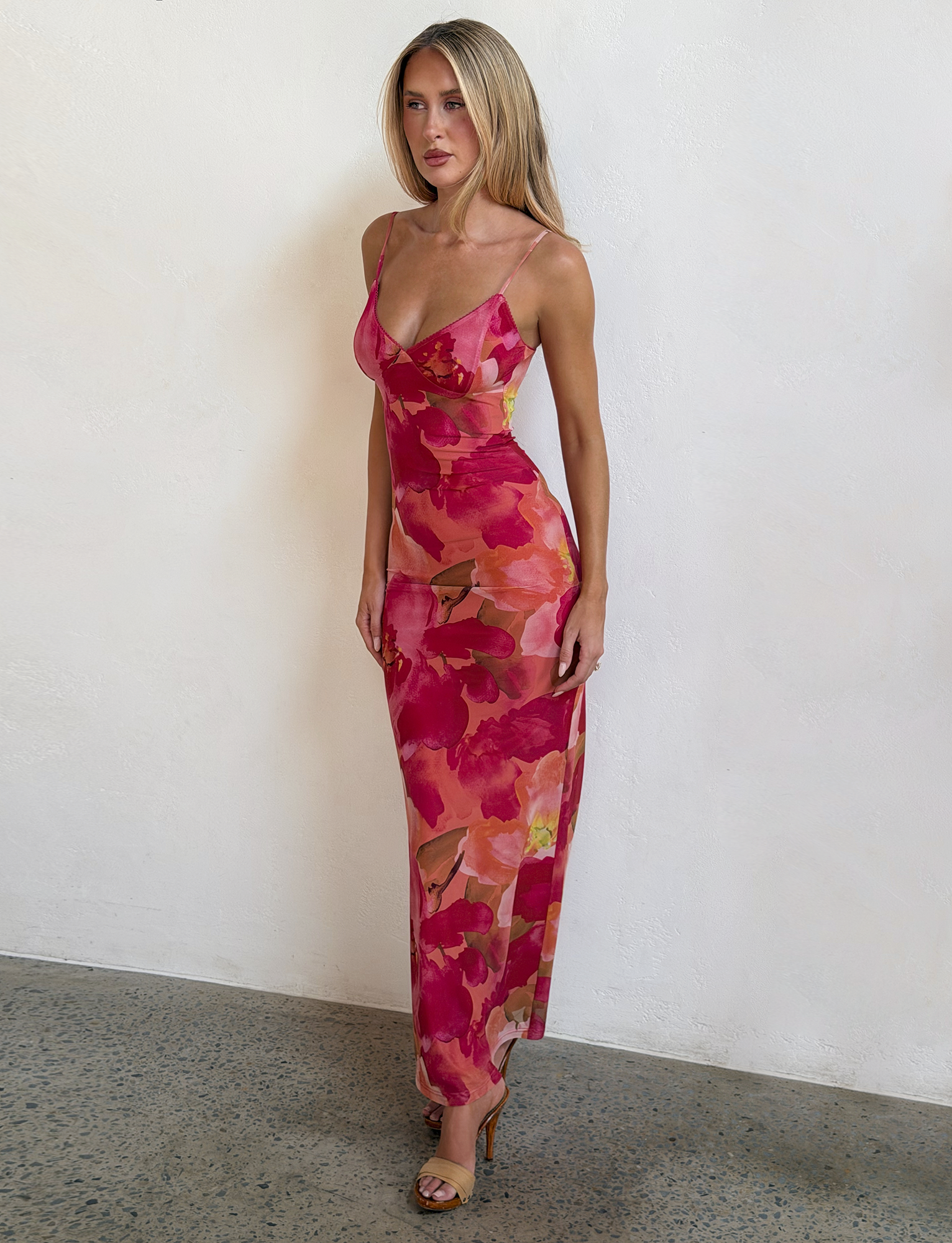 A woman with long straight hair and a neutral expression stands against a light wall, wearing the TIGERMIST JULIANNA MAXI DRESS - DAIQUIRI FLORAL with adjustable spaghetti straps and sandals.