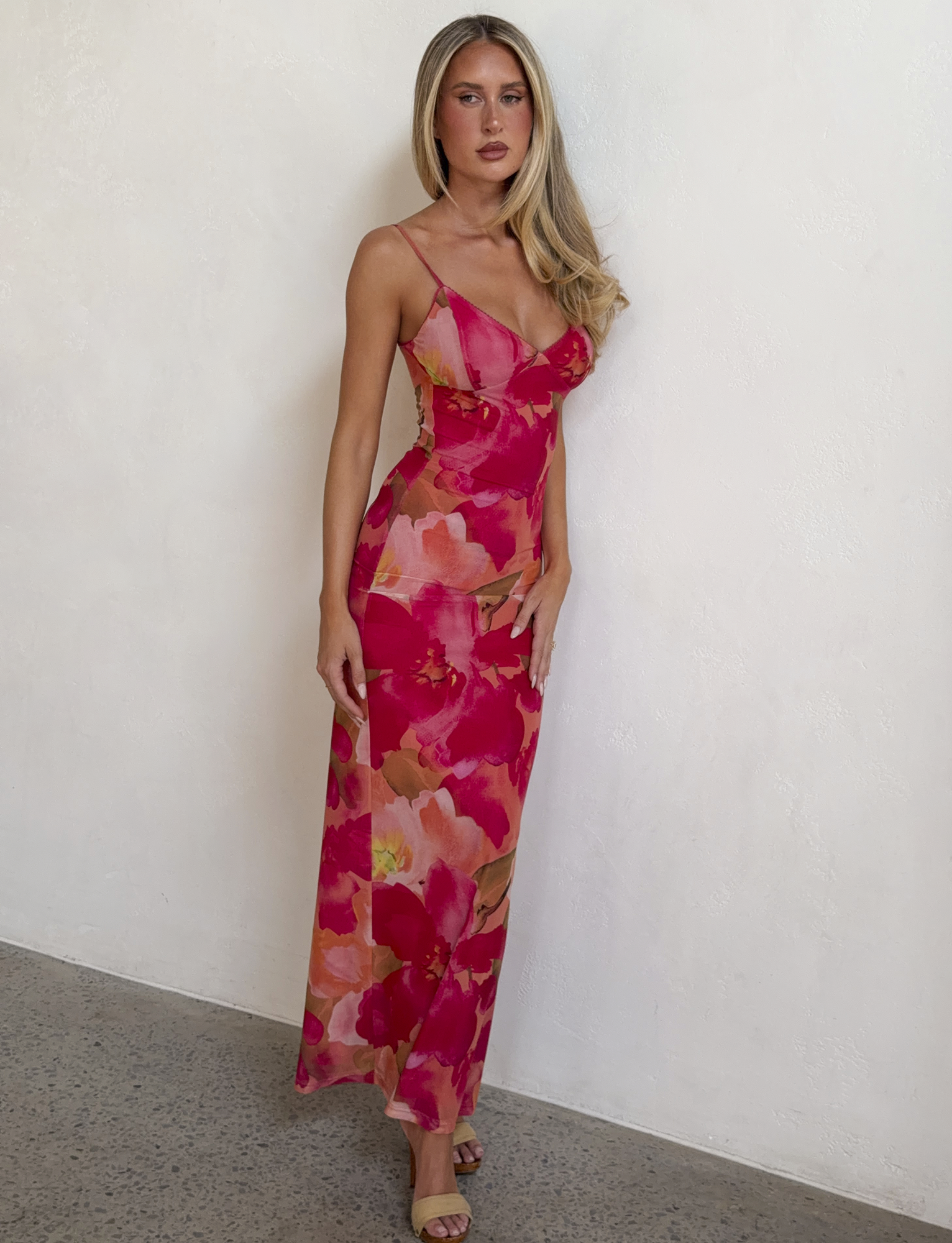 A woman with long blonde hair wears the TIGERMIST JULIANNA MAXI DRESS - DAIQUIRI FLORAL with adjustable spaghetti straps and beige heels, standing against a white textured wall and looking at the camera.