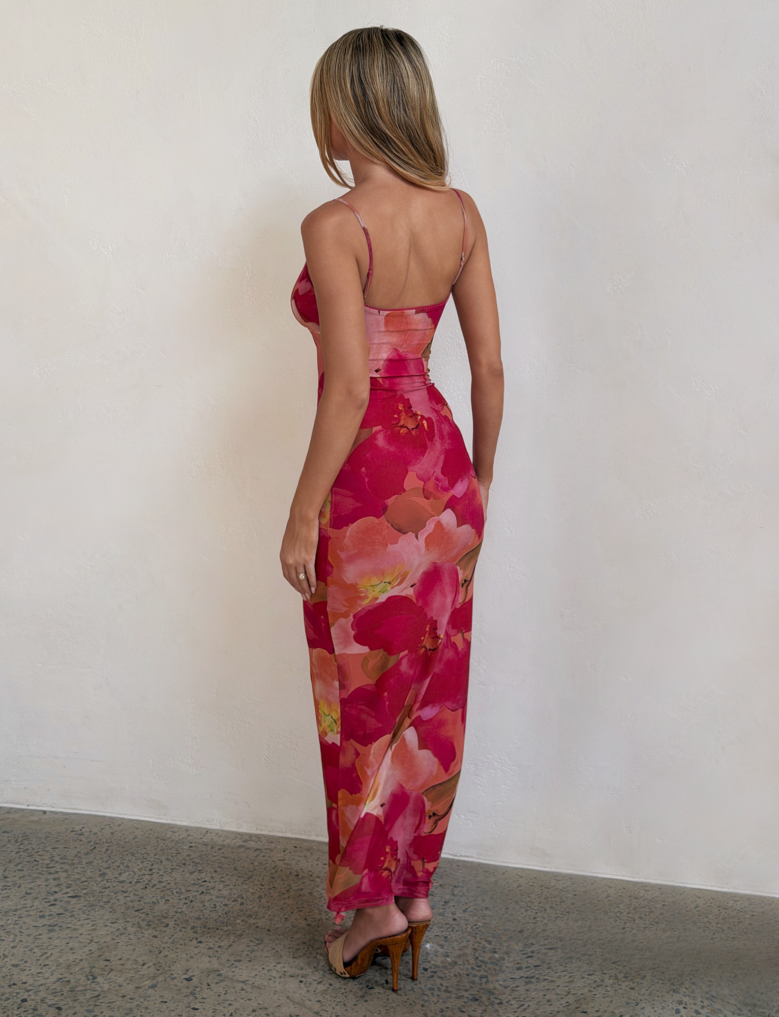 A woman stands indoors, back to camera, wearing the TIGERMIST JULIANNA MAXI DRESS - DAIQUIRI FLORAL with adjustable spaghetti straps and high heels against a light-colored textured wall.