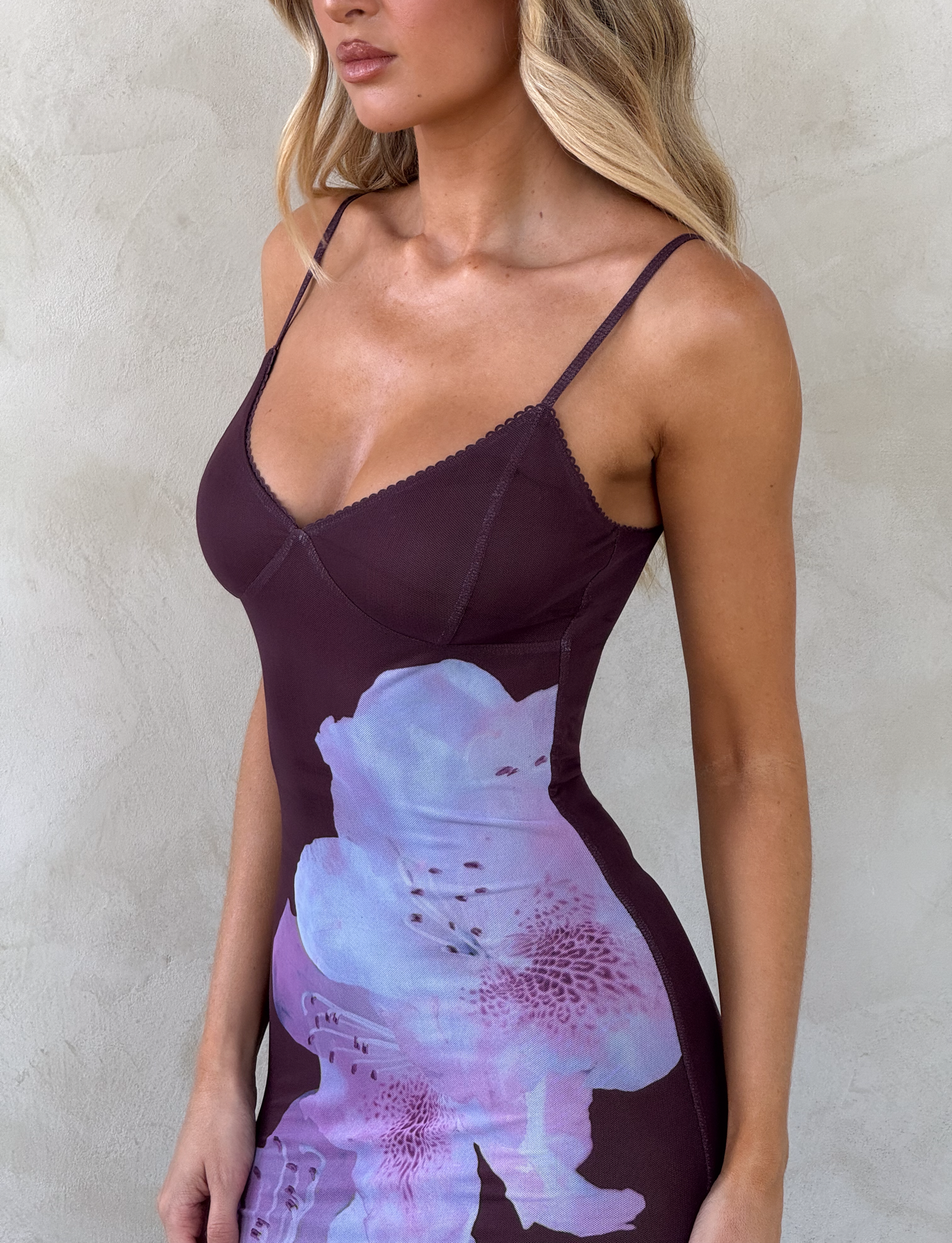 A woman models the TIGERMIST JULIANNA MAXI DRESS - ZEN FLORAL, featuring a fitted silhouette, thin straps, and a bold blue and purple floral print, while standing against a light-textured wall with her face partially out of frame.
