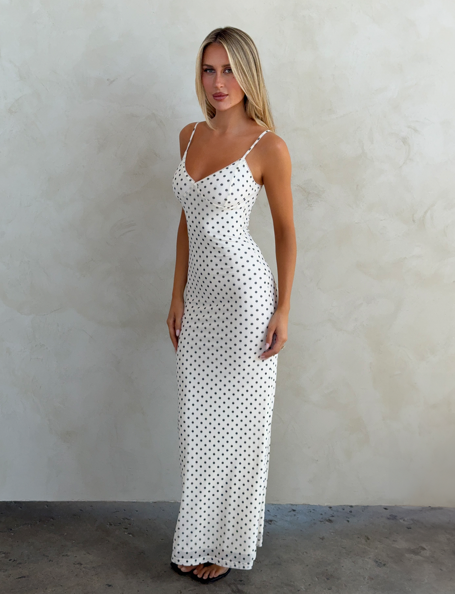 A woman with long blonde hair stands against a light textured wall, wearing the TIGERMIST JULIANNA MAXI DRESS - POLKA DOT, featuring black and white polka dots and adjustable spaghetti straps, hands resting at her sides.