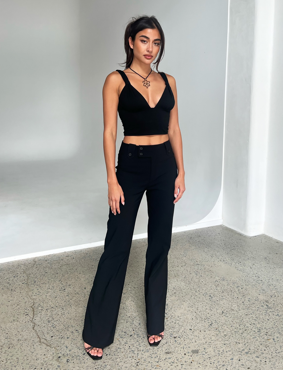 Indoors on a polished concrete floor, a woman with long dark hair pulled back wears TIGERMIST’s KITTIE HIGH RISE PANT in black—high-rise, flared, stretch-suiting fabric—paired with a black V-neck crop top, necklace, and black heels.