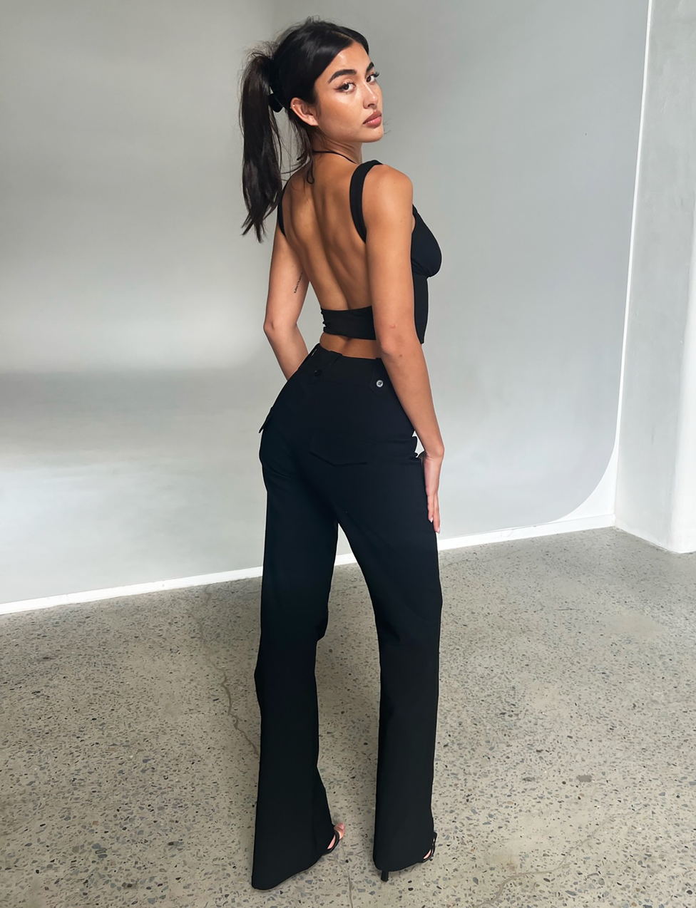 A woman with dark hair in a ponytail poses in a minimalist studio, wearing the TIGERMIST KITTIE HIGH RISE PANT - BLACK with a black backless top, standing on a speckled concrete floor and looking over her shoulder.