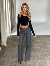 A woman with long blonde hair wears a black long-sleeve crop top and TIGERMIST's KITTIE HIGH RISE PANT - GREY with flared legs, standing in a studio setting with neutral curtains and a light backdrop.