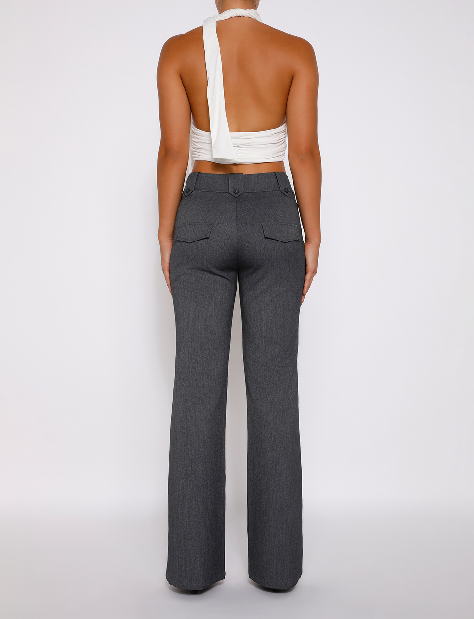 A person faces away, wearing TIGERMIST’s KITTIE HIGH RISE PANT - GREY featuring back pockets and a flared silhouette, paired with a sleeveless white asymmetrical wrap top against a plain white background.