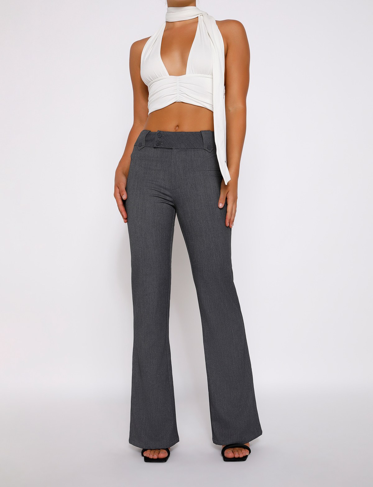 A woman stands against a plain white background wearing a sleeveless white halter crop top, TIGERMIST KITTIE HIGH RISE PANT in grey with flared legs, and black open-toe heels, her hands resting at her sides.