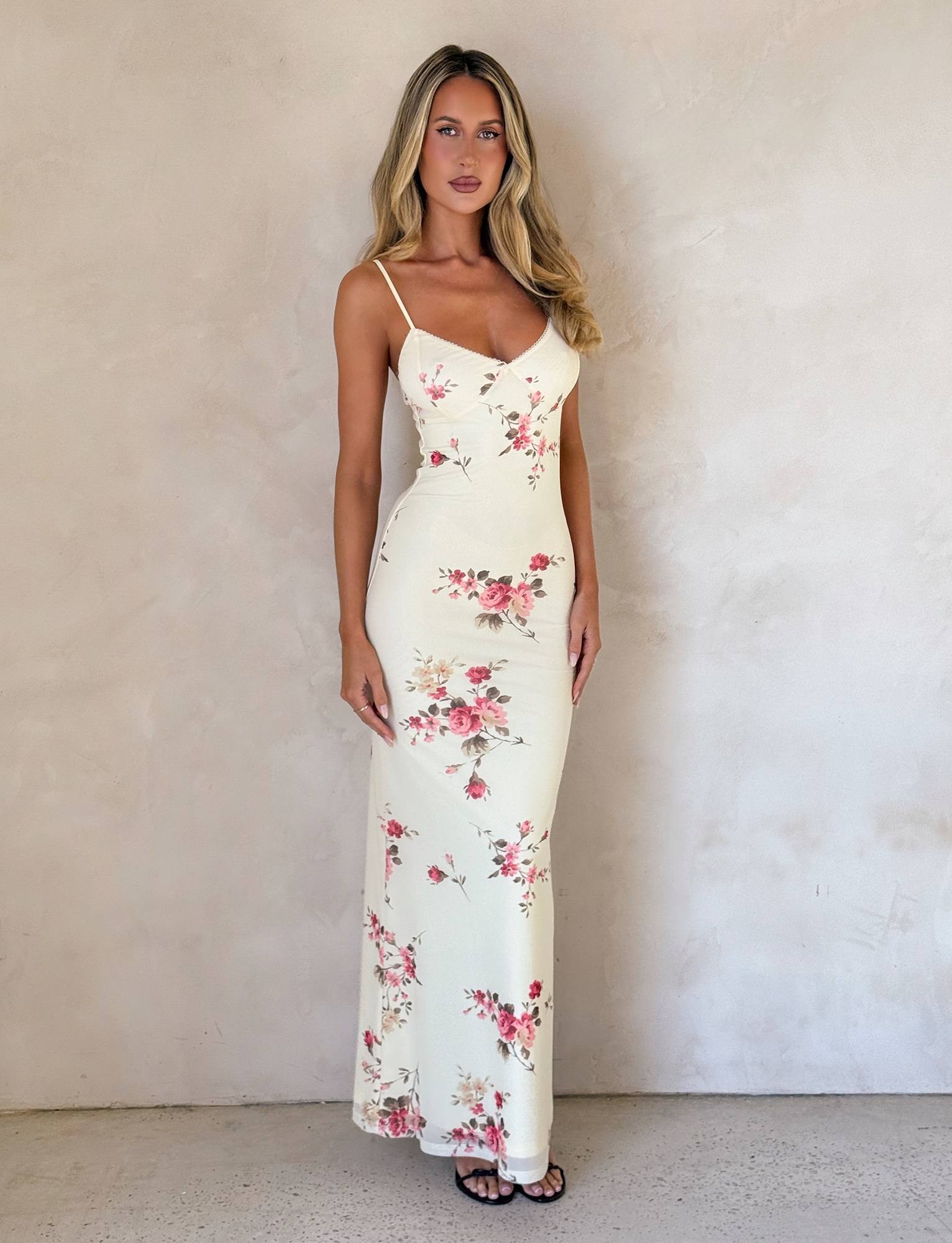 A woman with long blonde hair wears the TIGERMIST JULIANNA DRESS in yellow macro pop floral, featuring a fitted, sleeveless style and adjustable spaghetti straps, standing against a light textured wall and floor, looking at the camera.