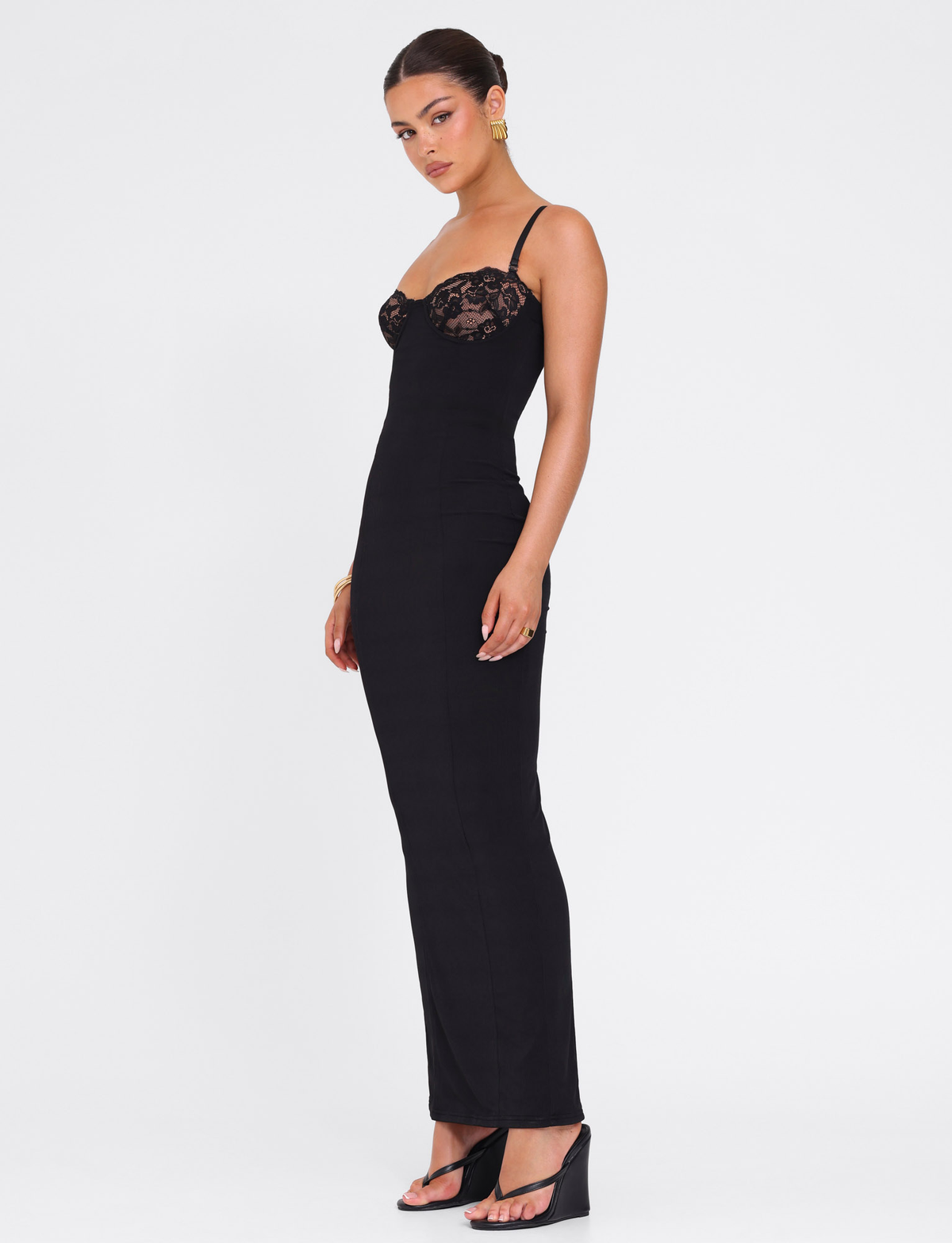 A woman stands against a white background wearing the TIGERMIST ALLURA MAXI DRESS in black, paired with black heels and gold earrings. Her hair is styled in a sleek bun, and she faces slightly to the side.