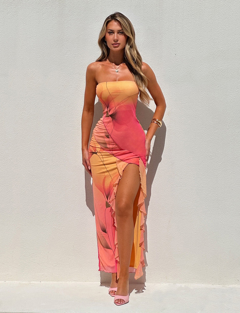 A woman with long, wavy hair stands against a white wall. She is wearing the stunning THEO MAXI DRESS - MULTI : MACRO FLORAL : GRADIENT FLORAL by TIGERMIST, featuring a strapless design in shades of pink, orange, and yellow with a thigh-high slit. She accessorizes with a necklace, bracelet, and strappy heels. The lighting creates soft shadows.