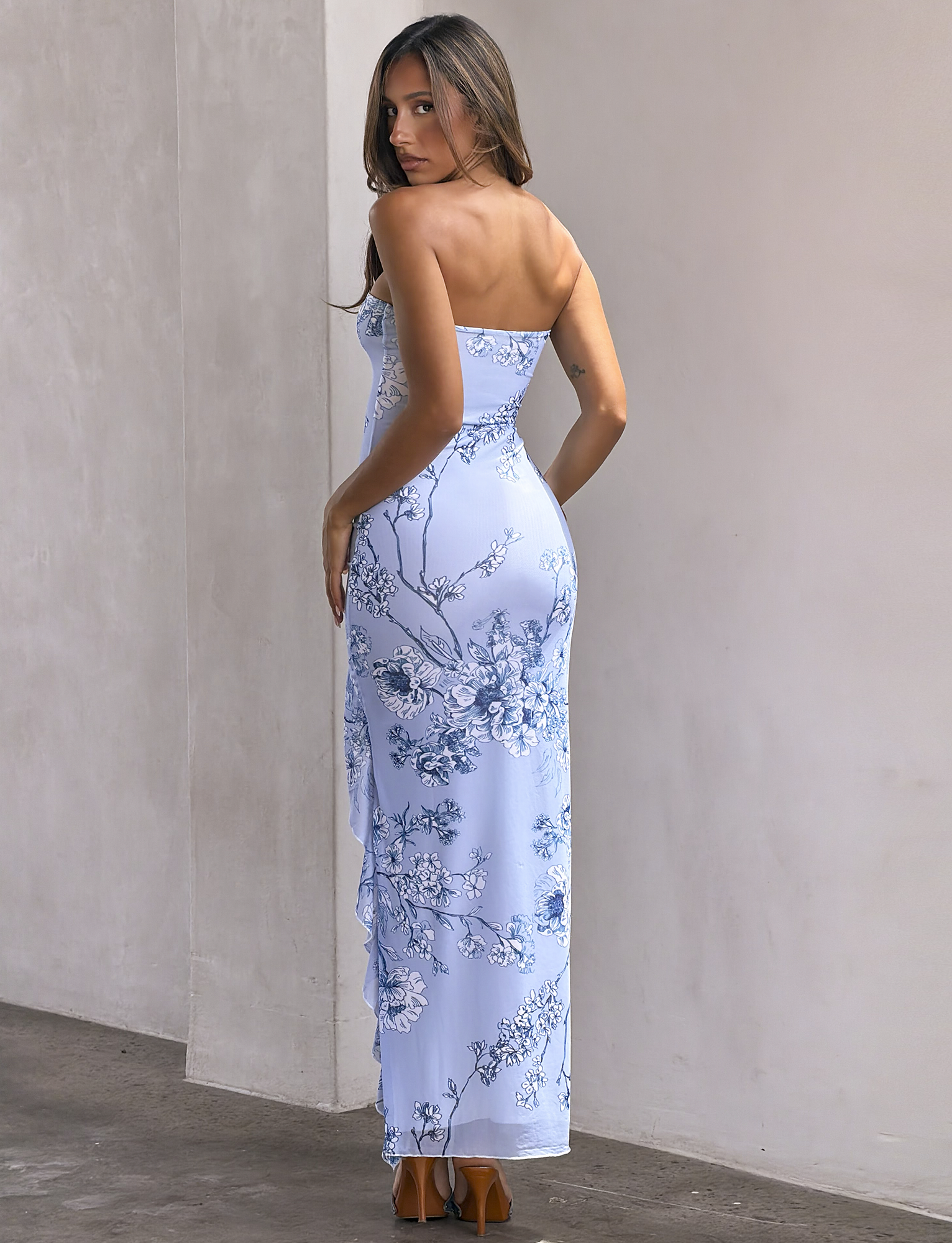 A woman faces away indoors, glancing over her shoulder at the camera. She wears the TIGERMIST THEO MAXI DRESS - BLUE FLORAL with brown heels, her long hair draped to one side against a plain, neutral background.