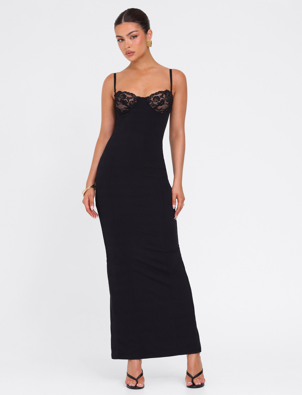 A woman poses in the TIGERMIST ALLURA MAXI DRESS - BLACK, a long, fitted bodycon dress with thin straps and lace bra cups. She pairs it with black heels, gold hoop earrings, and a bracelet against a plain white background.