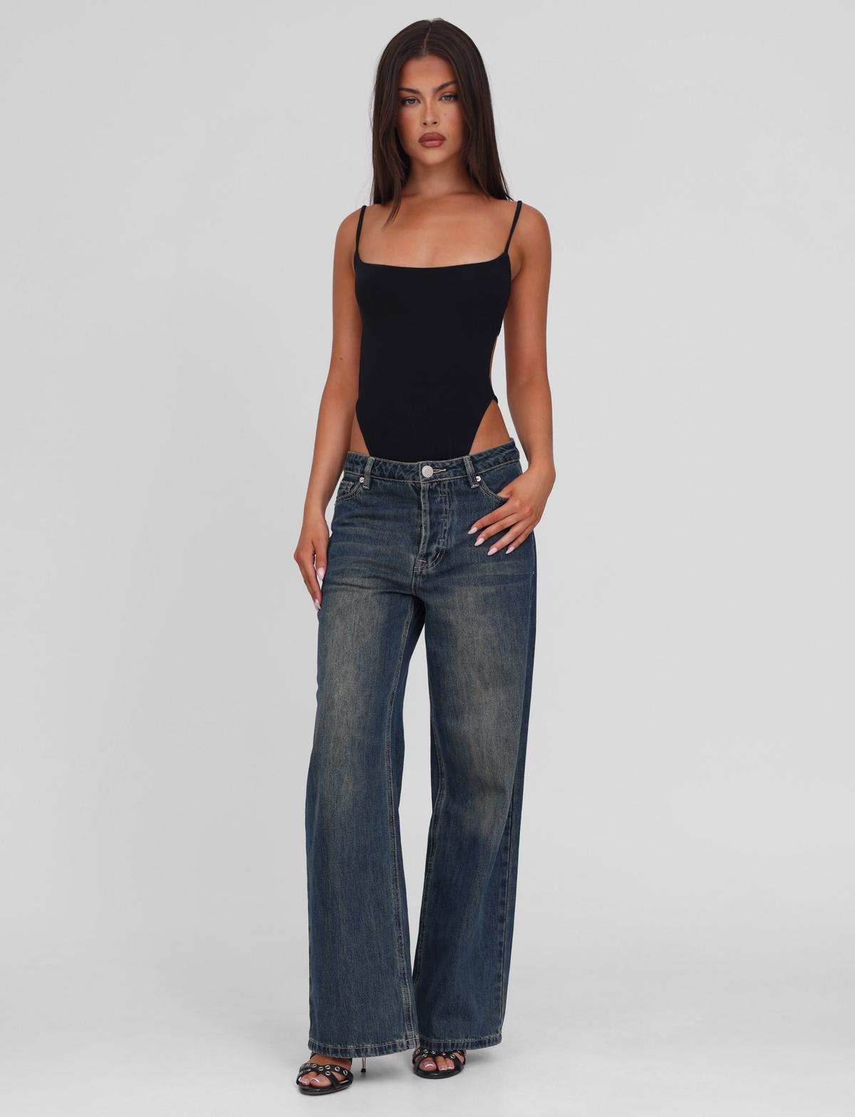 A woman with long brown hair models the TIGERMIST MINERVA BODYSUIT in black, styled with loose blue jeans. She stands against a light gray background, looking forward with a neutral expression.