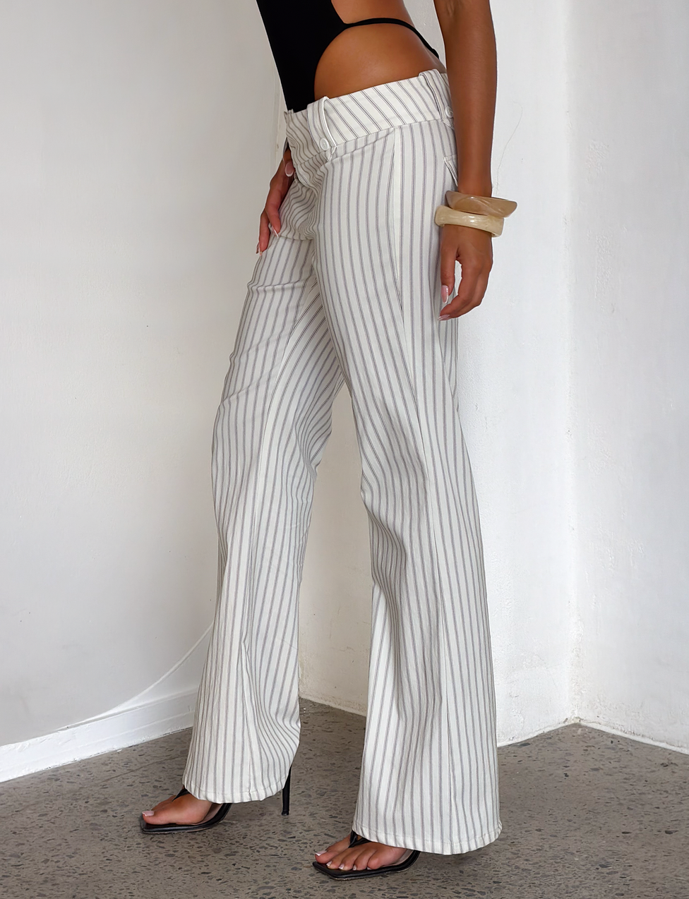 A person wears TIGERMIST’s KITTIE PANT - IVORY STRIPE, styled with a black top, black heels, and two chunky bangles. Only the lower body is visible as they stand against a white wall.