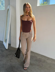 A woman with long blonde hair wears a maroon fitted top and TIGERMIST KITTIE PANT in taupe with flared legs, carrying a black patterned handbag. She stands indoors near a mirror and sunlit windows, looking to the side.