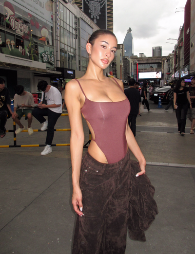 A woman wears the TIGERMIST MINERVA BODYSUIT in chocolate with dark brown pants, posing confidently on a city street lined with busy shops and tall buildings beneath a cloudy sky.