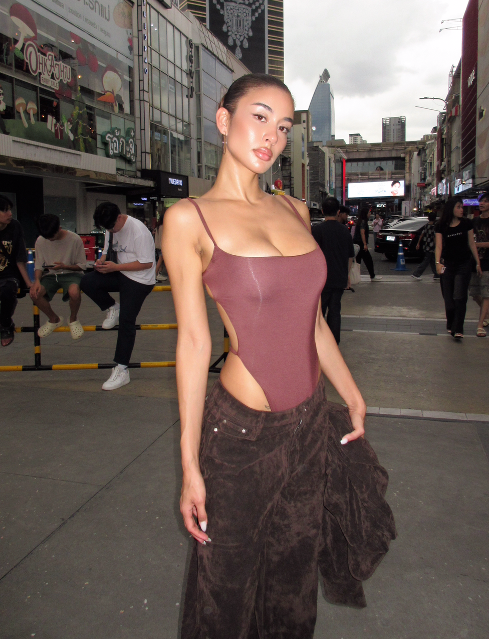 A woman wears the TIGERMIST MINERVA BODYSUIT in chocolate with dark brown pants, posing confidently on a city street lined with busy shops and tall buildings beneath a cloudy sky.