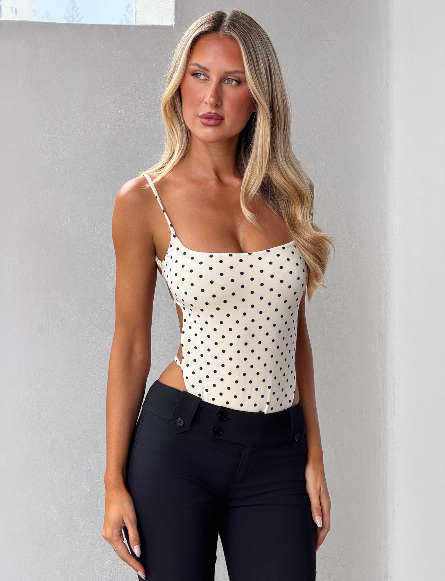 A woman with long blonde hair wears the CL - MINERVA BODYSUIT by Tiger Mist USA, a white, sleeveless, polka dot top, paired with black pants. She stands indoors against a white wall and looks to the side.