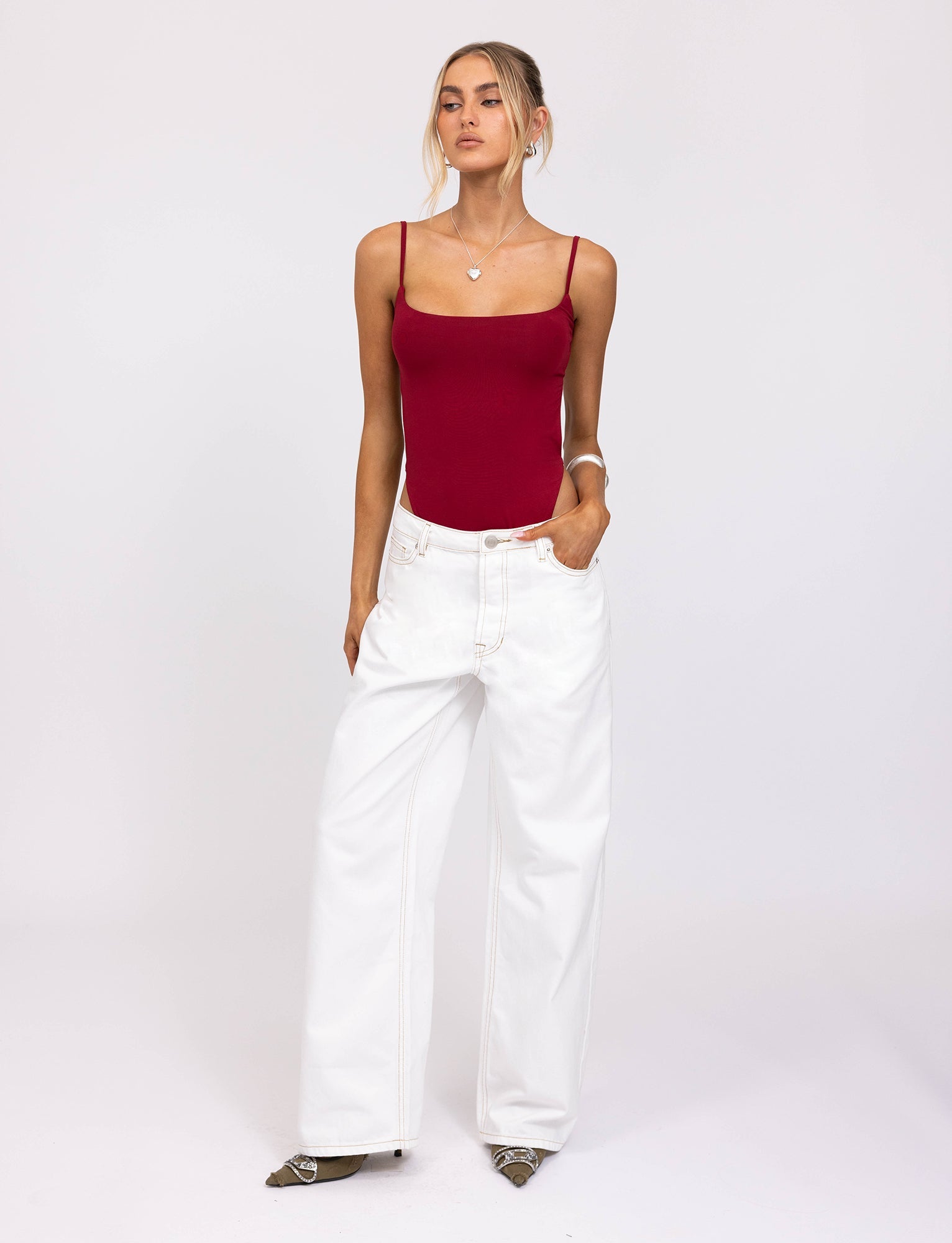 A person with light-colored hair styled in a loose updo is wearing the fitted, sleeveless MINERVA BODYSUIT - RED from TIGERMIST, paired with high-waisted, wide-leg white pants. With one hand in their pocket, they stand against a plain white background.