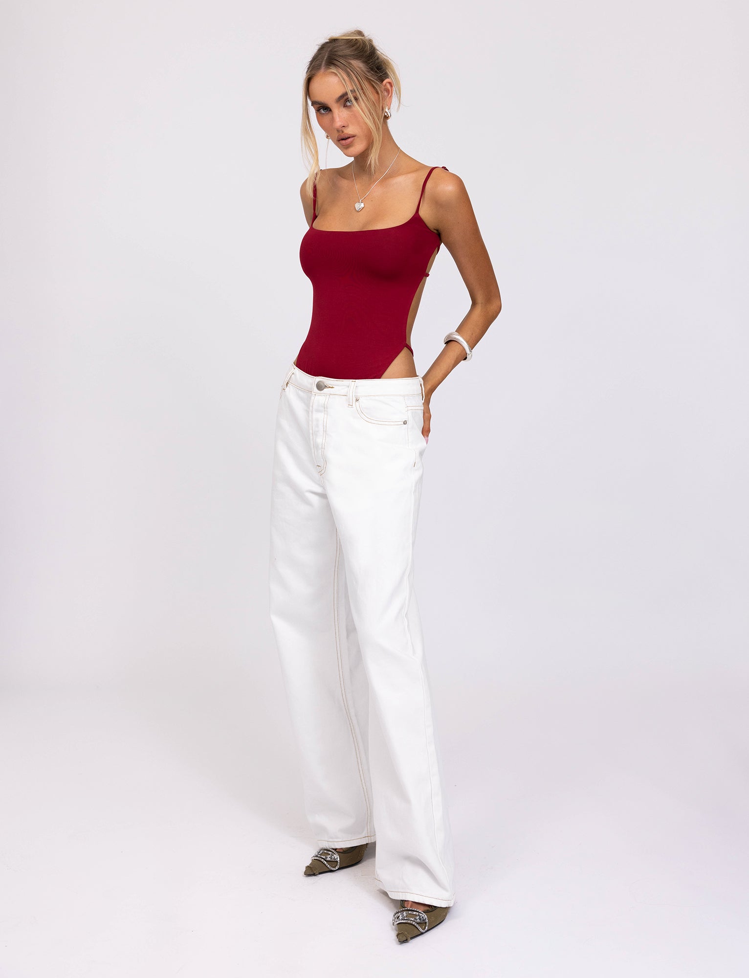 A woman with long hair stands against a plain background. She is wearing the MINERVA BODYSUIT in red from TIGERMIST, paired with high-waisted white wide-leg pants. One hand in her pocket and the other resting on her hip, she accessorizes with a necklace and bracelet.