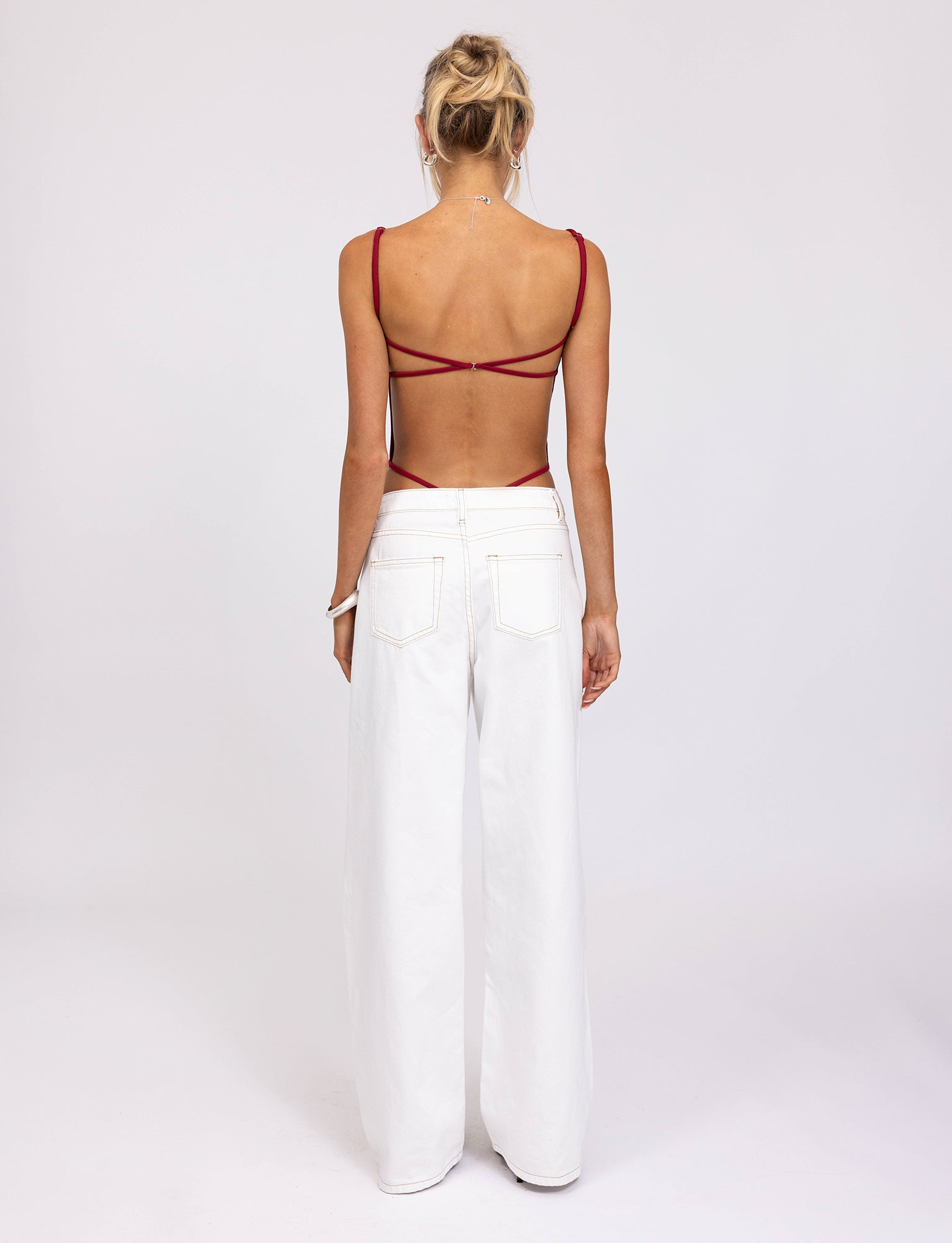 A person with blonde hair tied in a bun is facing away from the camera, wearing a red MINERVA BODYSUIT from TIGERMIST and high-waisted white wide-leg pants with pockets. Their outfit, reminiscent of Tiger Mist's chic aesthetic, stands out against the plain white background.