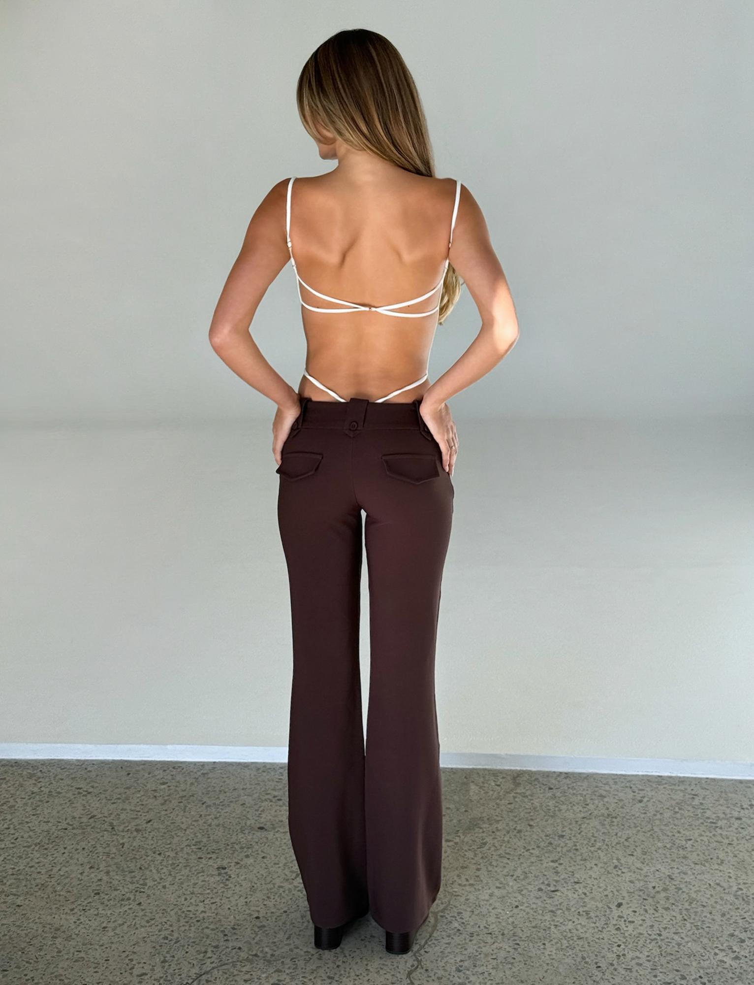 A woman with long, straight hair stands with her back to the camera, hands on hips, wearing TIGERMIST's MINERVA BODYSUIT in white and high-waisted brown flared pants.