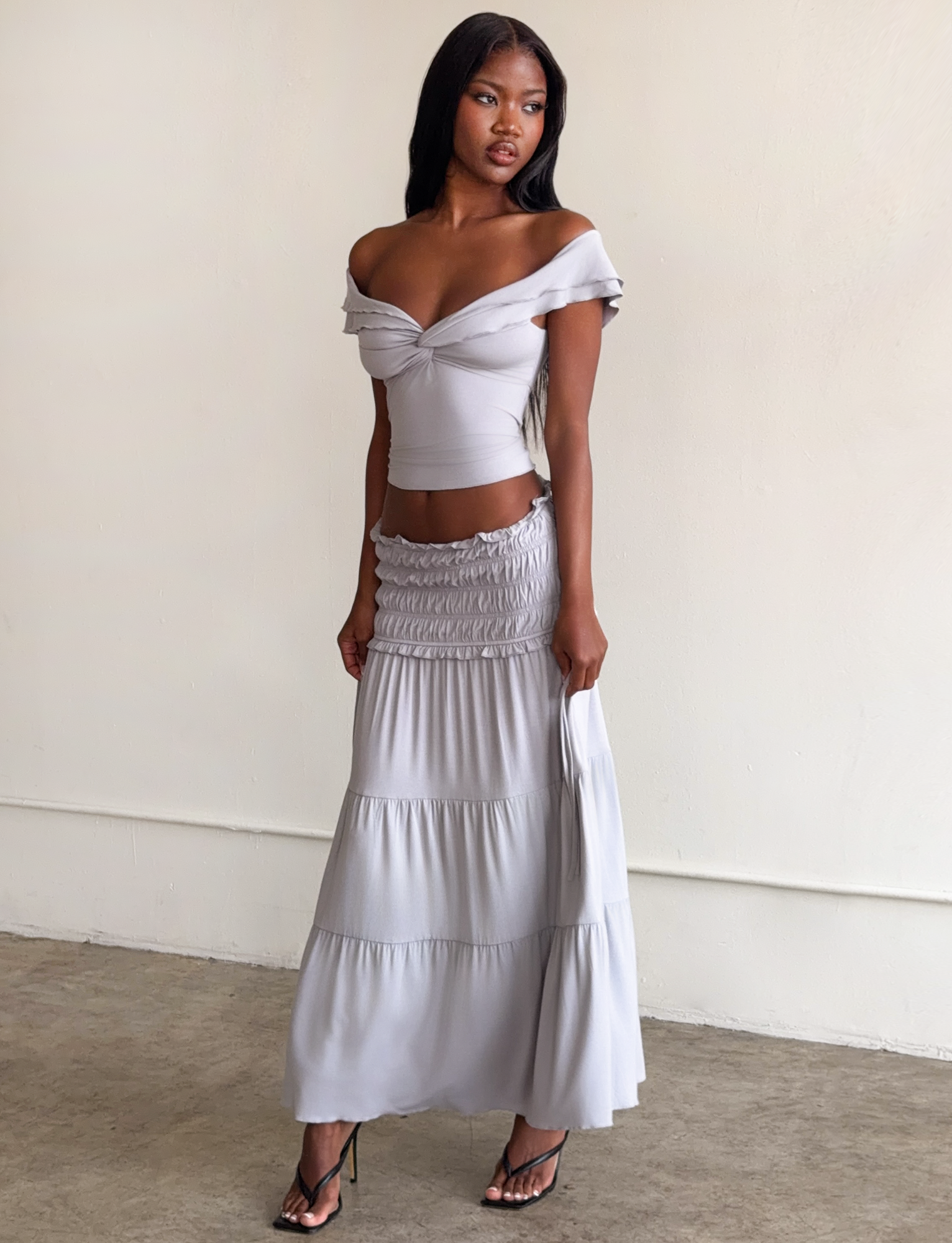 A woman stands indoors in the TIGERMIST DENALI MAXI SKIRT in dove grey, featuring a shirred waistband. She pairs it with a light gray off-the-shoulder crop top and strappy high-heeled sandals. The background is plain and minimal.